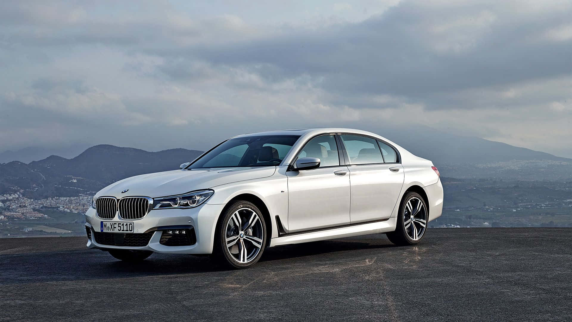 Bmw 7 Series 桌布 壁紙 — 一輛時尚的銀色寶馬轎車停在鋪砌的觀景點上，背景是遠山和陰天。