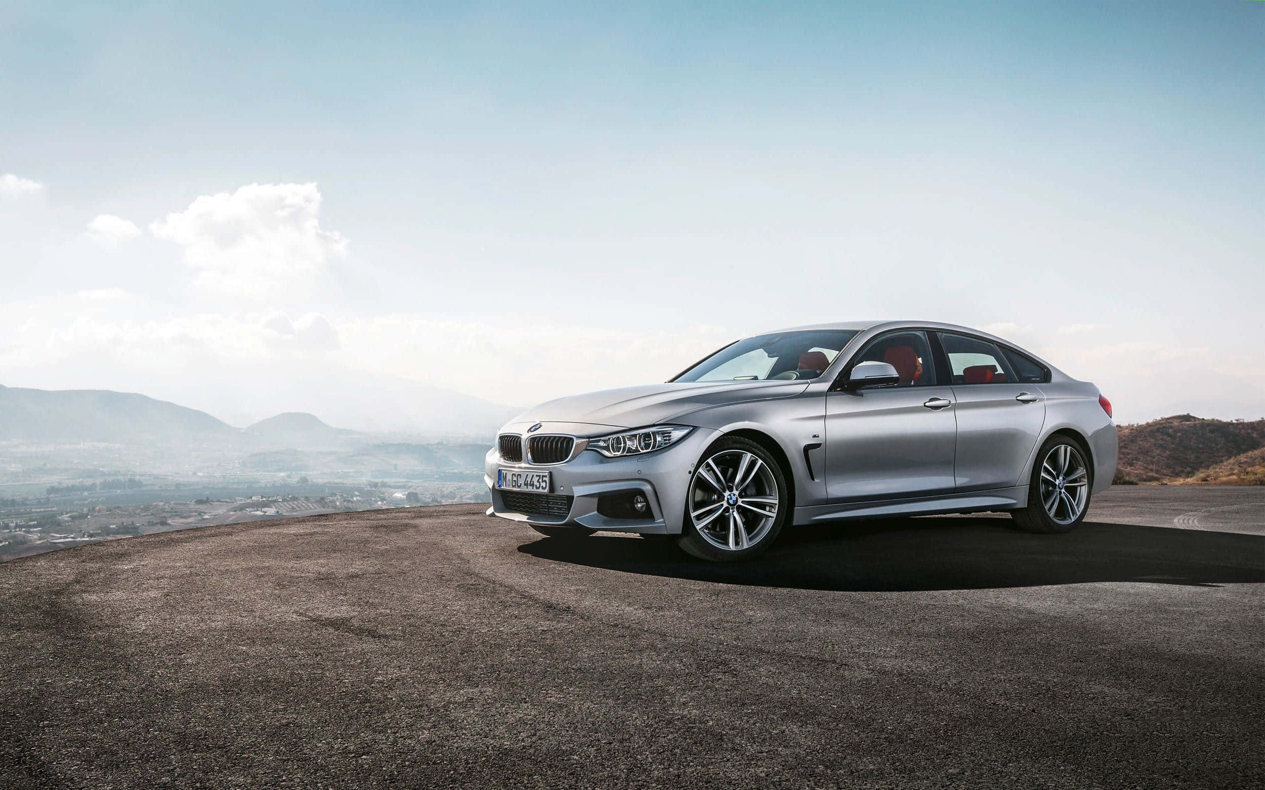 Bmw 4 Series 桌布 壁紙 — 一輛時尚的銀色寶馬轎車停在高處觀景點，遠山和朦朧的城市天際線在明亮天空下。