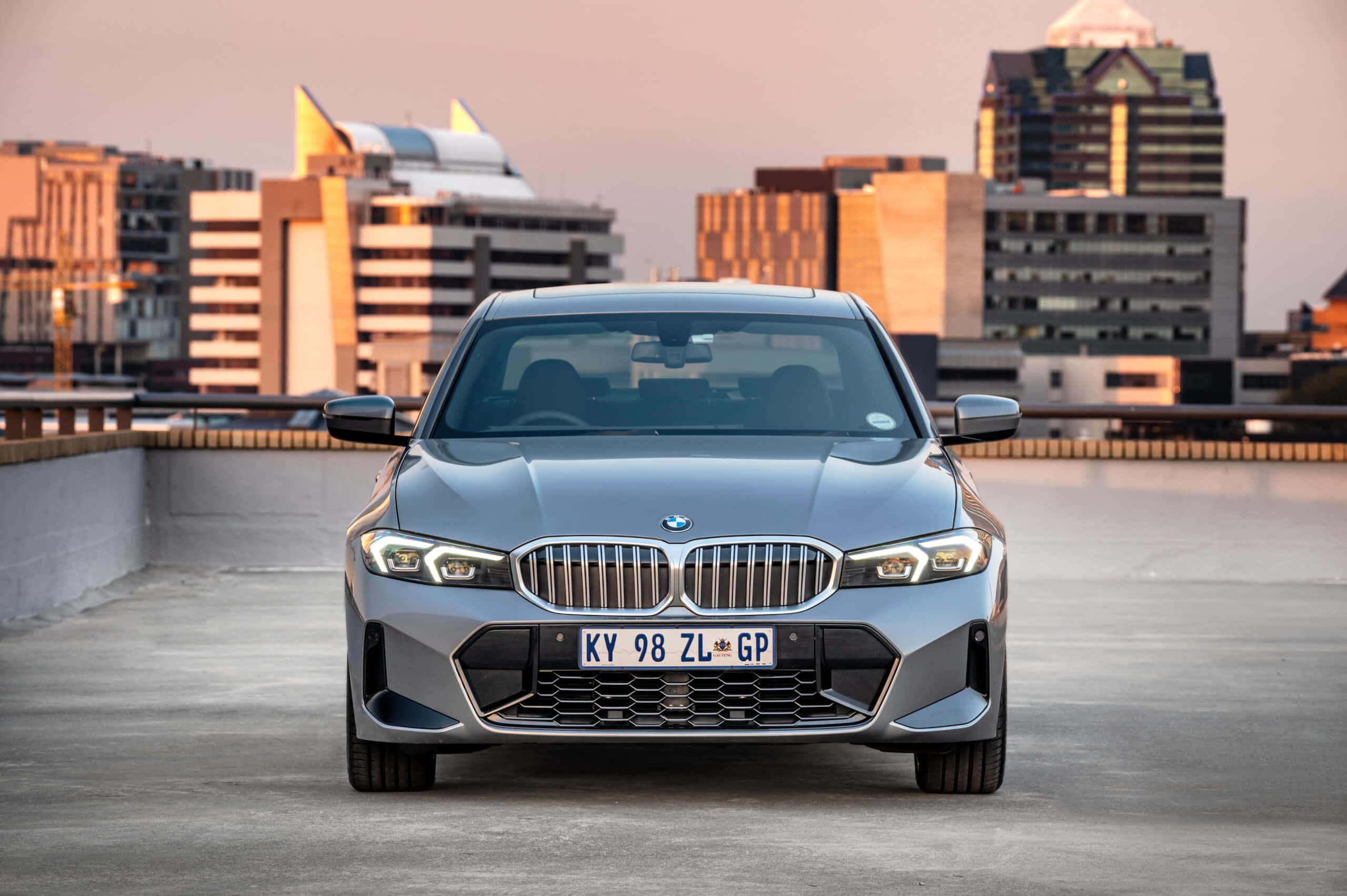 Bmw 3 Series 桌布 壁紙 — 一輛時尚的銀色寶馬轎車在屋頂上朝前，背後是溫暖夕陽光輝映照的城市天際線。