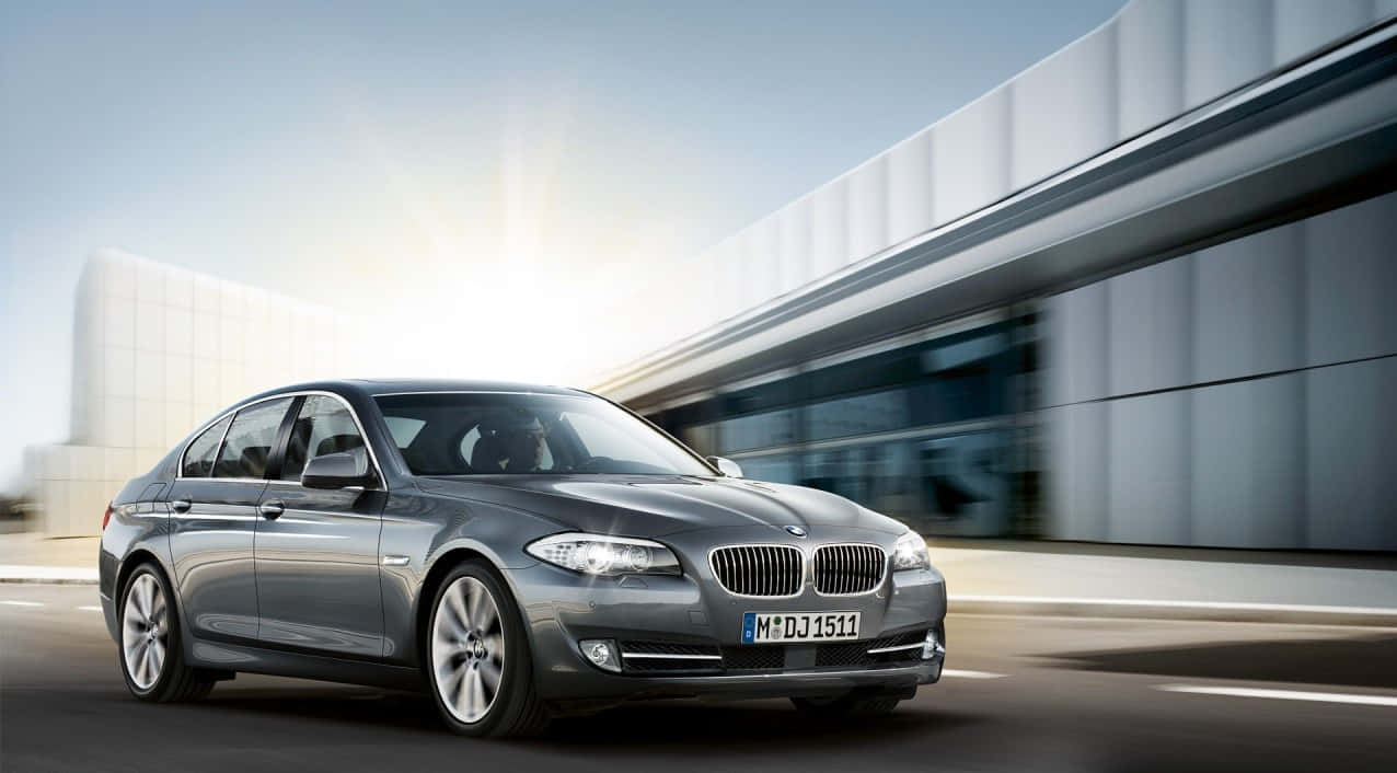 BMW 動圖 桌布 壁紙 — 一輛時尚的銀色寶馬轎車在明亮的陽光下飛馳而過現代玻璃建築。
