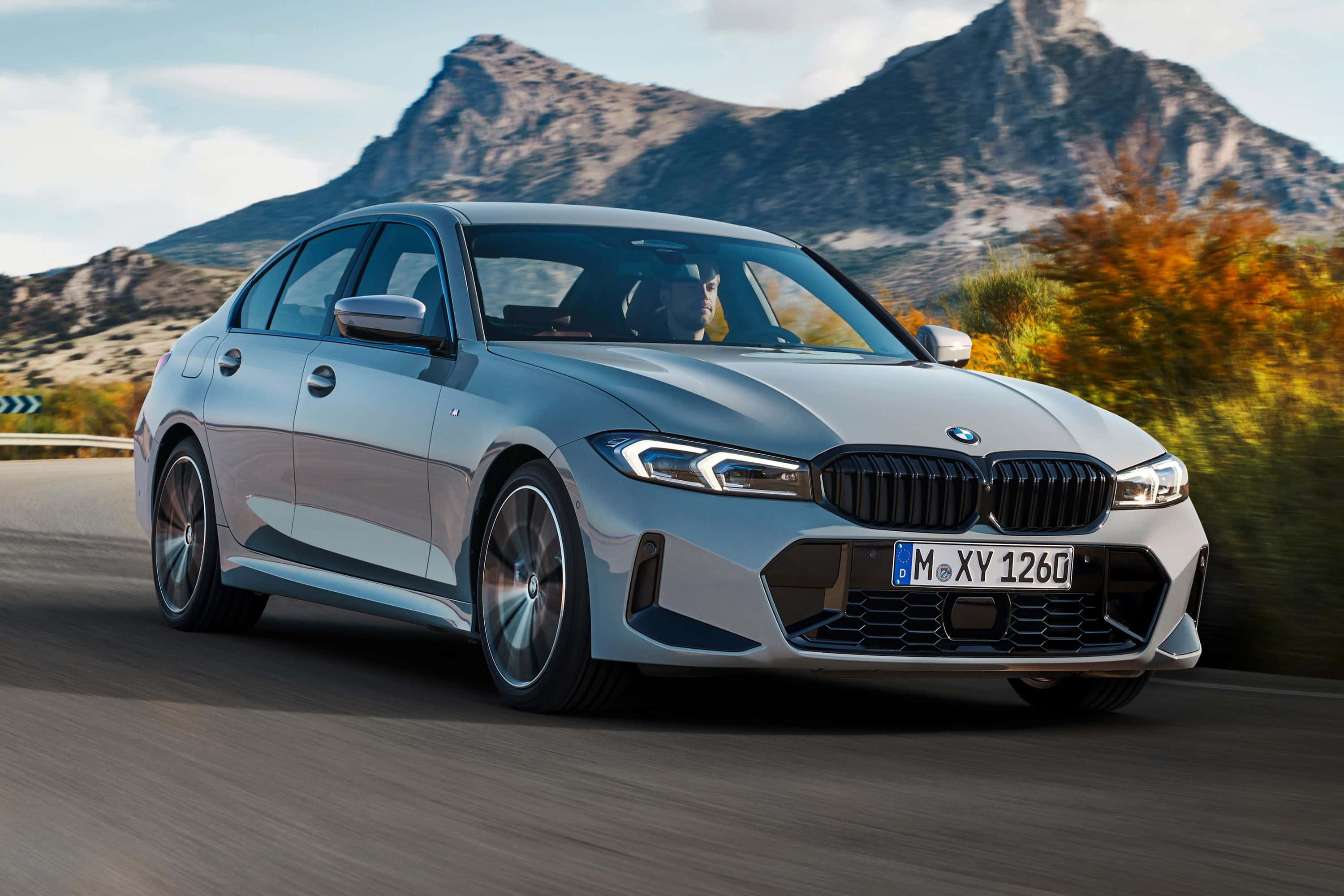 Bmw 3 Series 桌布 壁紙 — 一輛時尚的銀色寶馬轎車在秋葉和崎嶇山峰背景下沿山路疾馳。