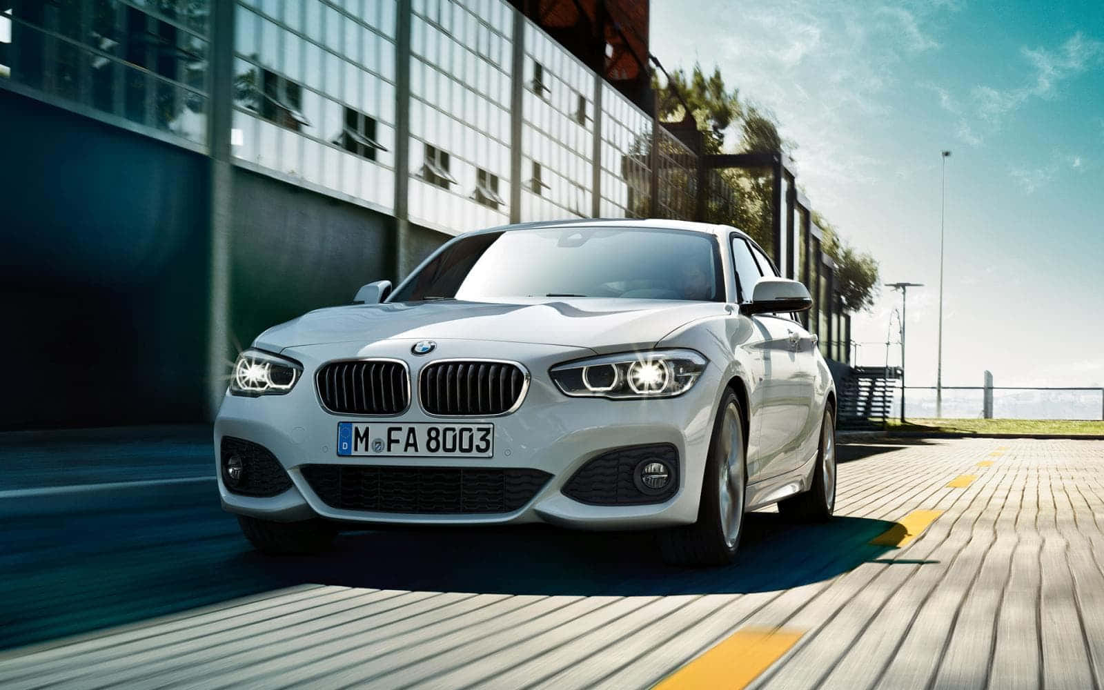 Bmw 1 Series 桌布 壁紙 — 一輛時尚的銀色寶馬轎車在陽光普照的城市道路上疾馳，背景是現代化的玻璃建築。