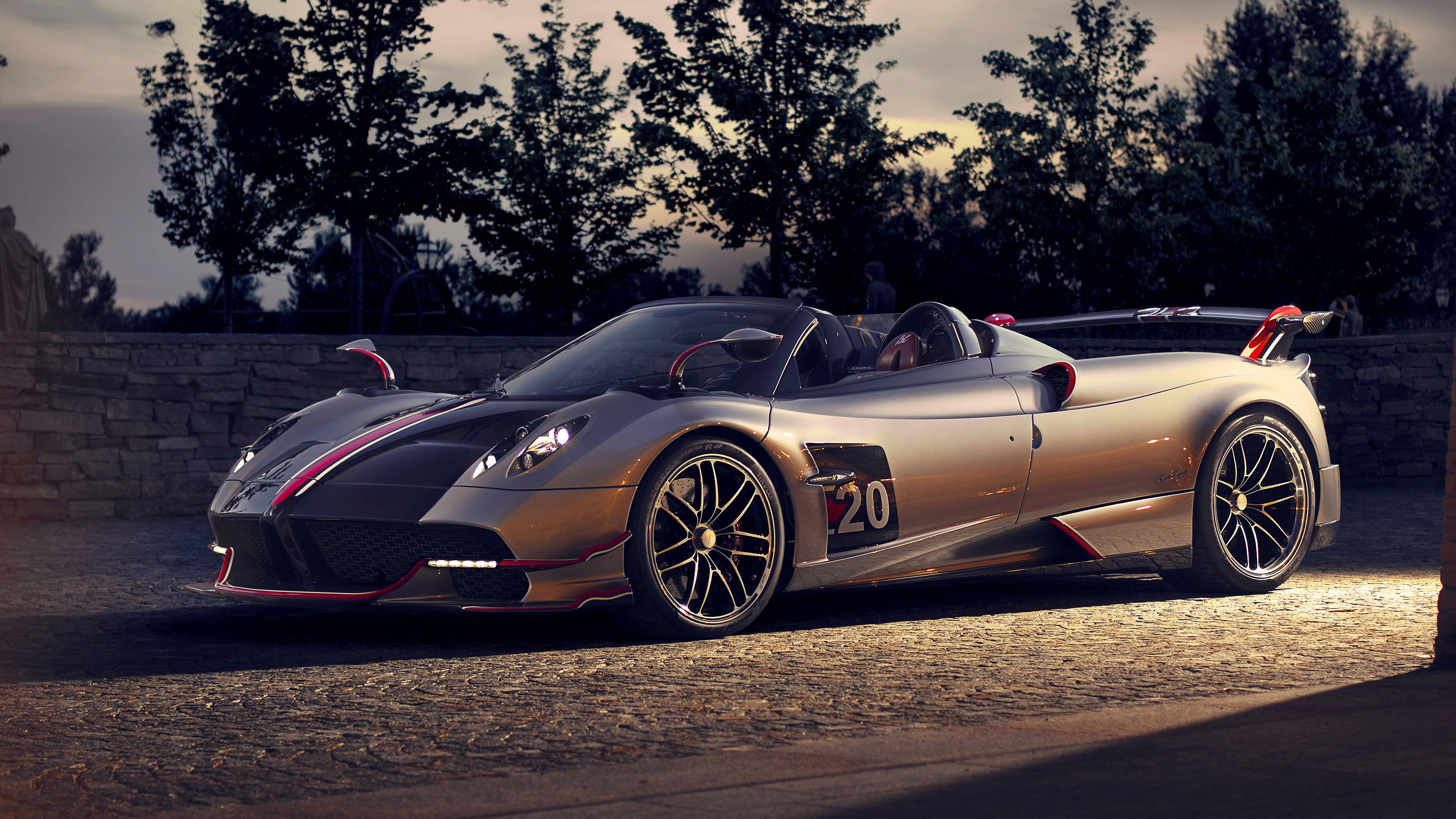 Pagani 桌布 壁紙 — 一輛時尚的銀色帕加尼Huayra Roadster，帶有紅色點綴，停泊在黃昏的鵝卵石路上，背景有樹木。