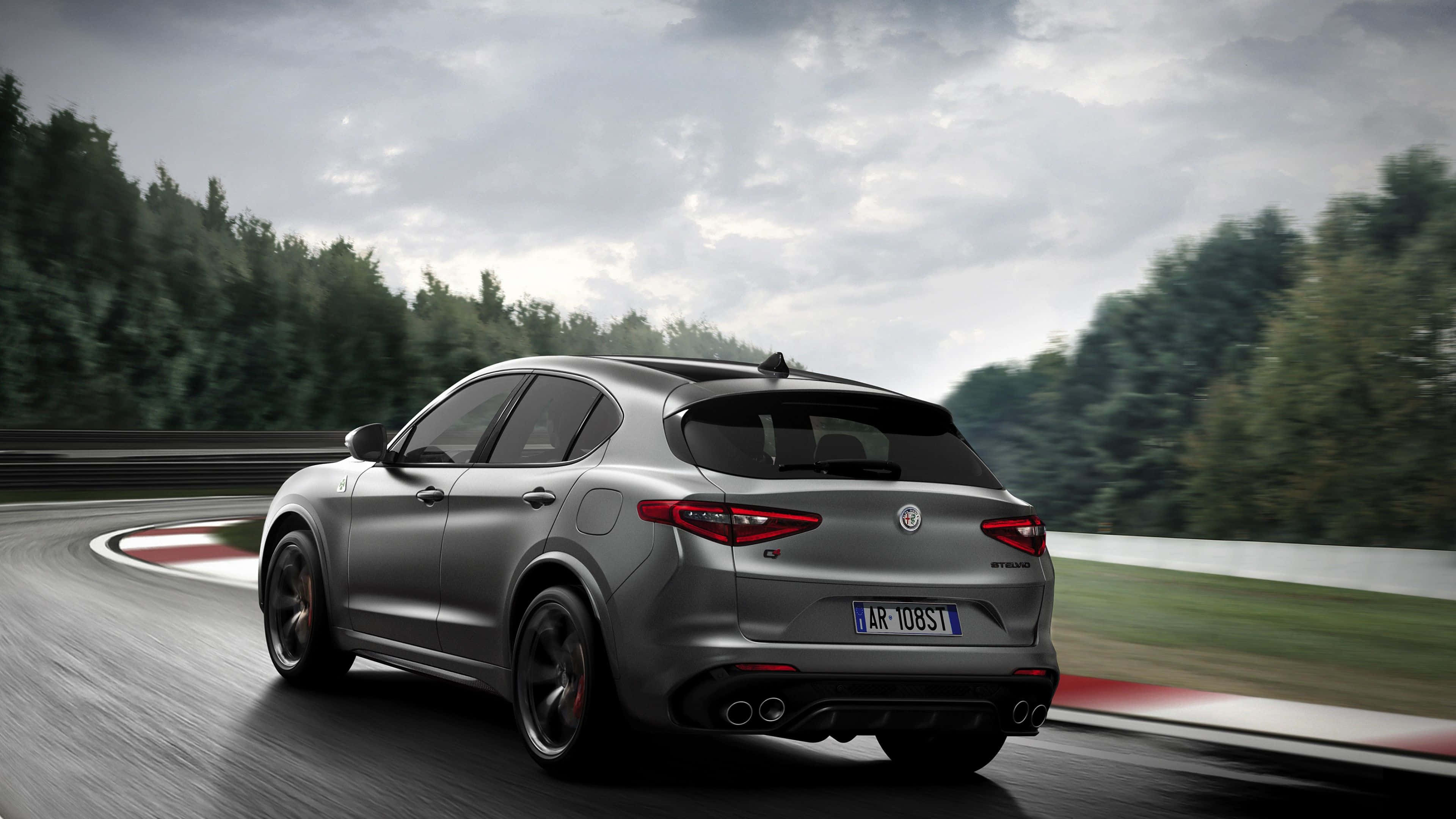 Alfa Romeo Stelvio 桌布 壁紙 — 一輛時尚的銀色愛快羅密歐 Stelvio Quadrifoglio 在潮濕的賽車場彎道上飛馳，背景是模糊的綠色樹木。