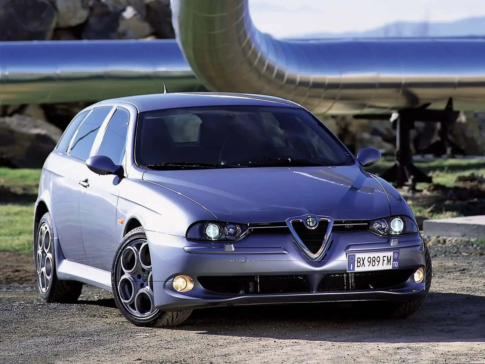 Alfa Romeo 156 桌布 壁紙 — 一輛時尚的銀色愛快羅密歐147跑車停在碎石上，背景有大型管道和岩石。