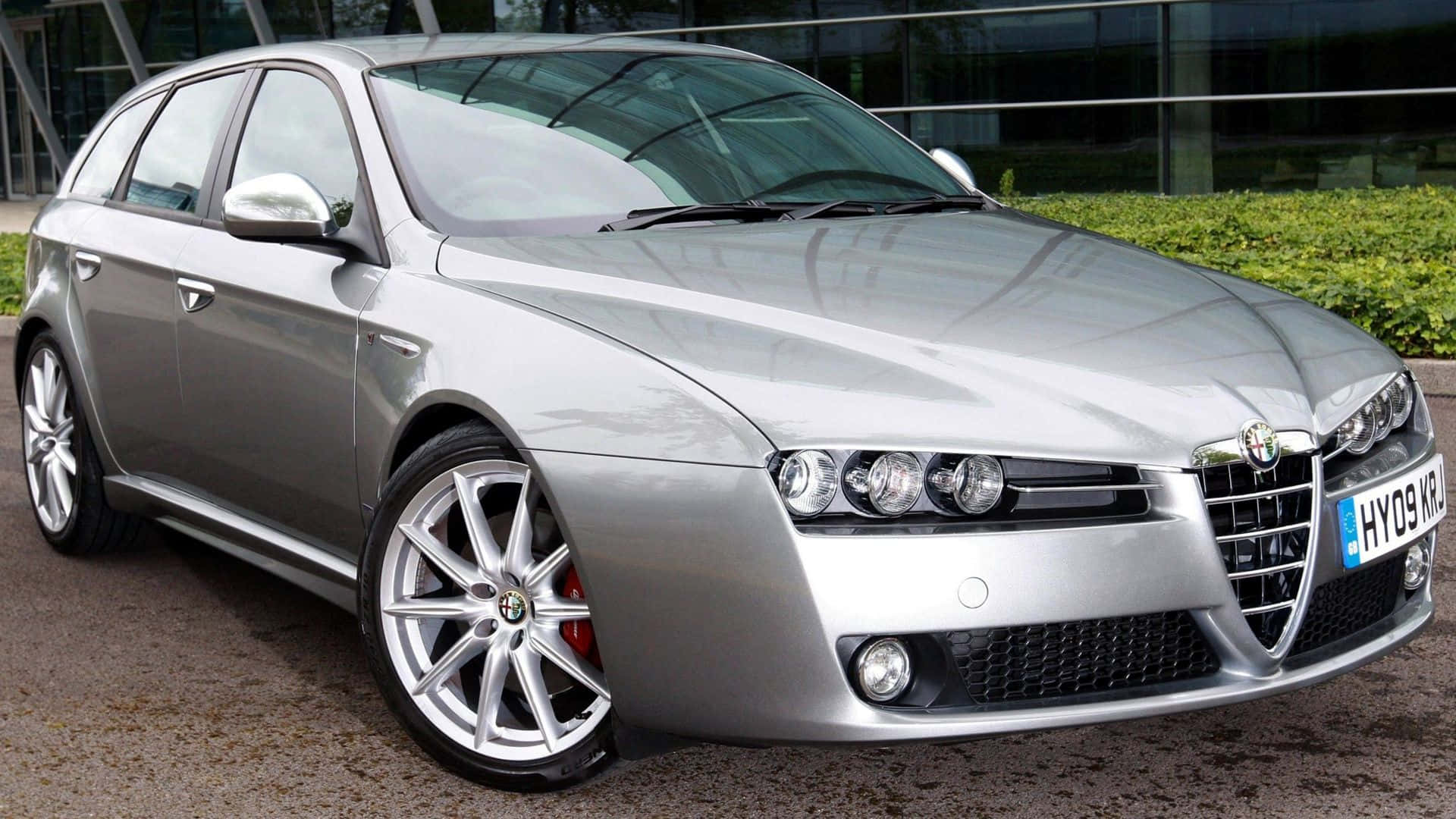 Alfa Romeo 159 桌布 壁紙 — 一輛時尚的銀色愛快羅密歐旅行車停在碎石路上，背景是現代玻璃建築和綠色樹籬。