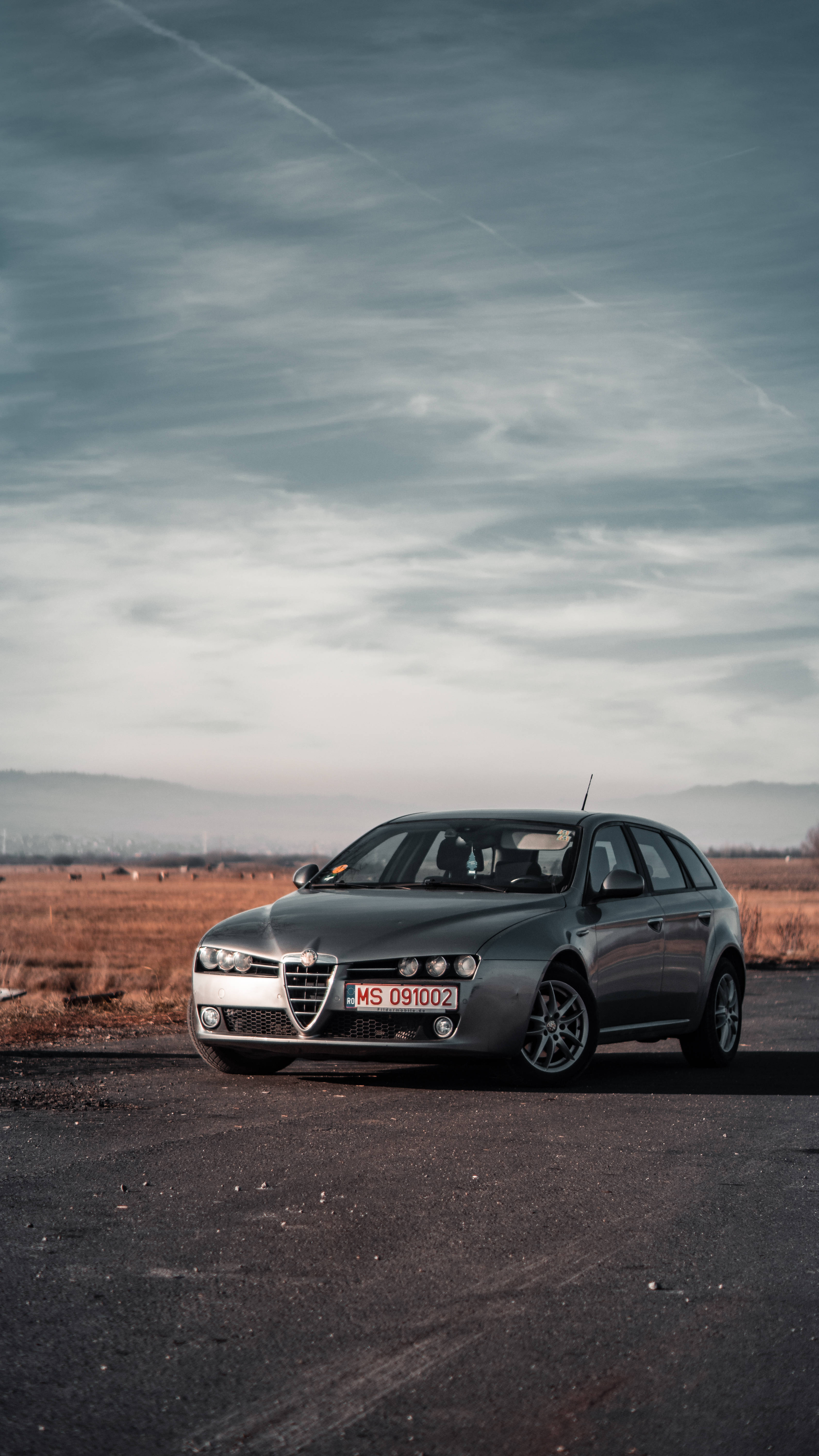 alfa romeo iPhone 桌布 壁紙 — 一輛時尚的銀色愛快羅密歐旅行車停泊在瀝青路上，背景是廣闊多雲的天空和遠山。