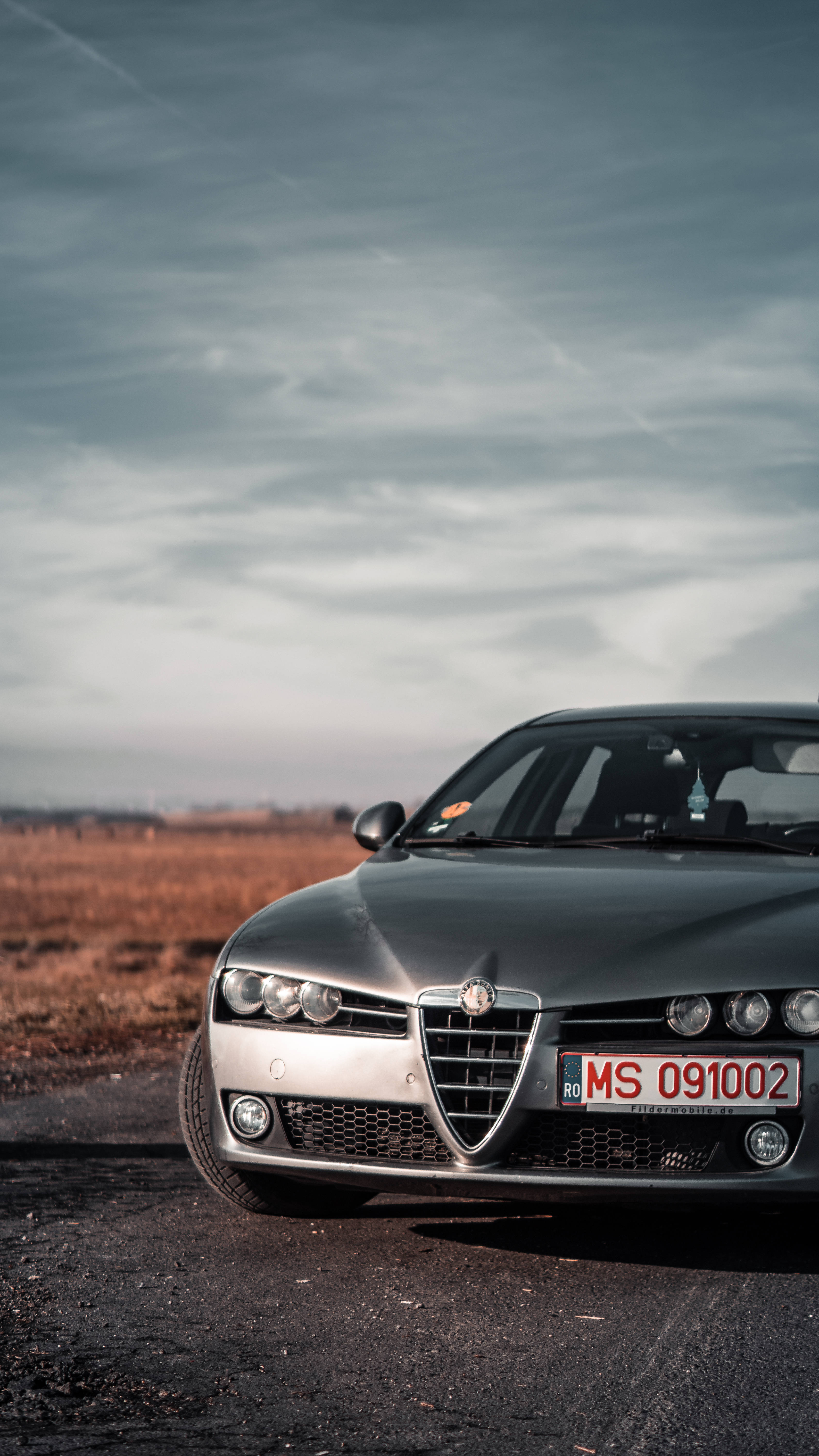 alfa romeo iPhone 桌布 壁紙 — 一輛時尚的銀色愛快羅密歐跑車停泊在碎石路上，天空多雲，背景有乾草。