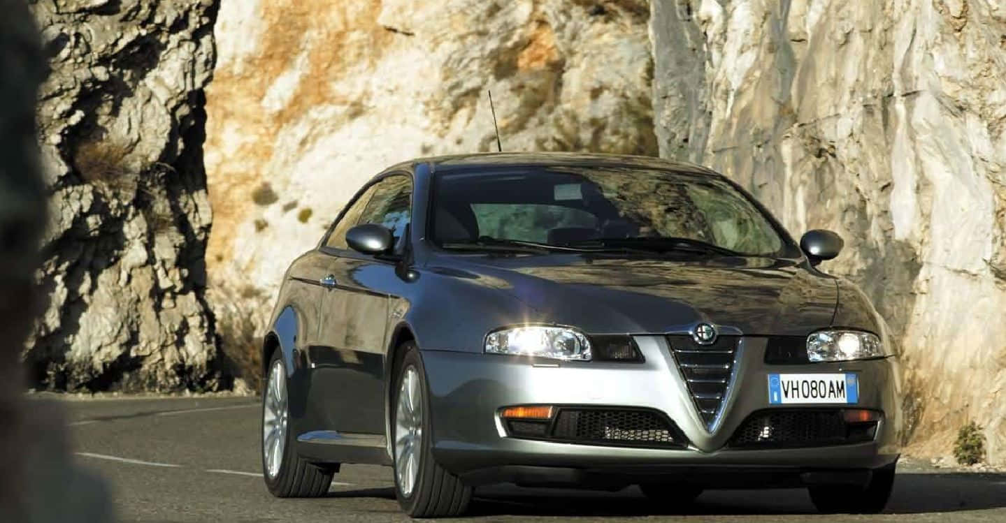 Alfa Romeo Gt 桌布 壁紙 — 一輛時尚的銀色愛快羅密歐轎跑車在明亮的日光下行駛在蜿蜒的山路上，兩旁是崎嶇的岩石峭壁。