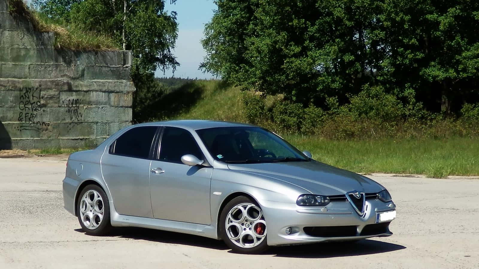 Alfa Romeo 156 桌布 壁紙 — 一輛時尚的銀色愛快羅密歐轎車停放在混凝土停車場，旁邊是長滿青苔的石牆和翠綠樹木，在明亮的日光下。