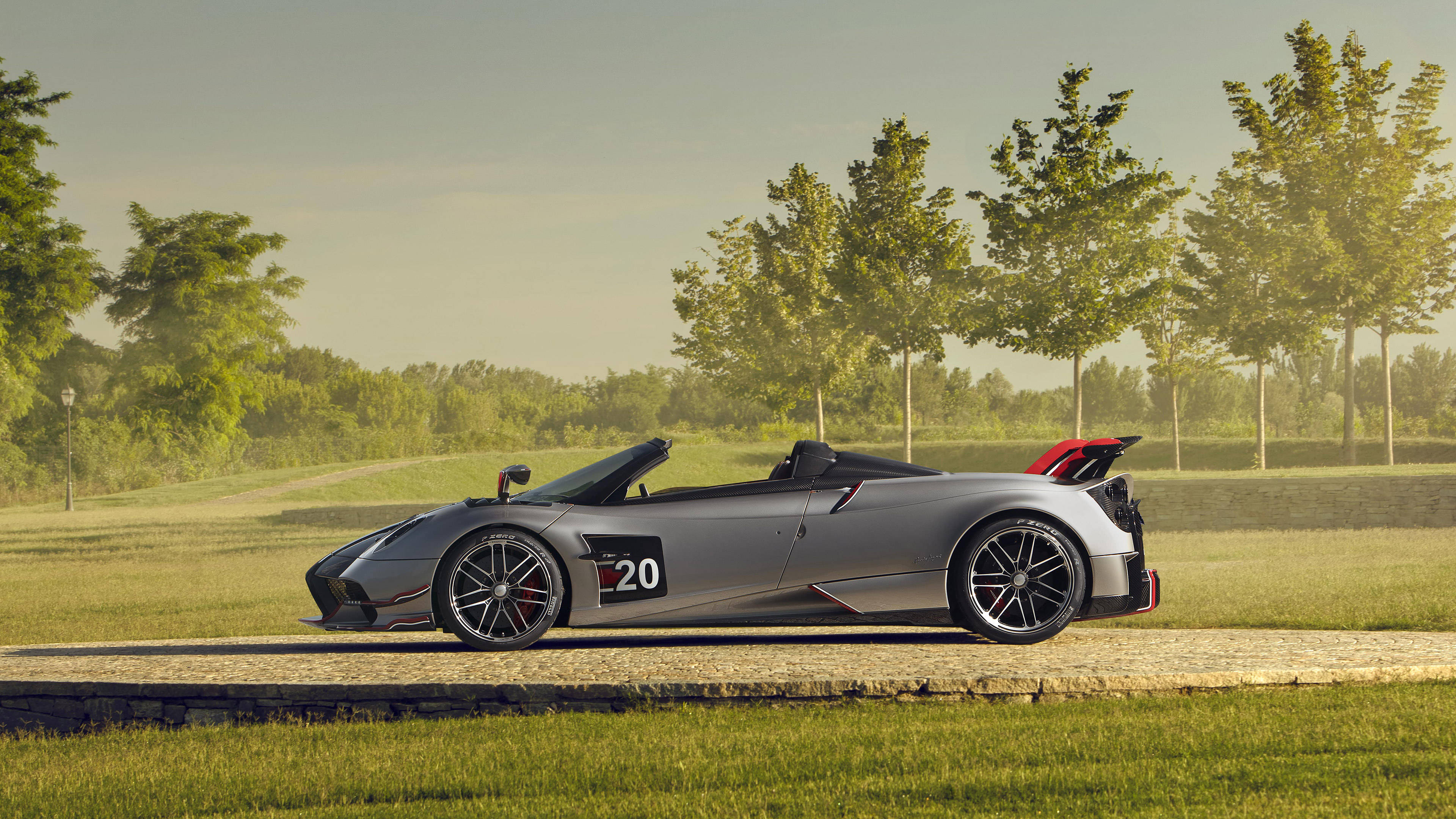 pagani iPhone 桌布 壁紙 — 一輛時尚的銀色敞篷跑車，帶有賽車號碼20，停在碎石路上，旁邊是草地和樹木，沐浴在柔和的金色光線中。