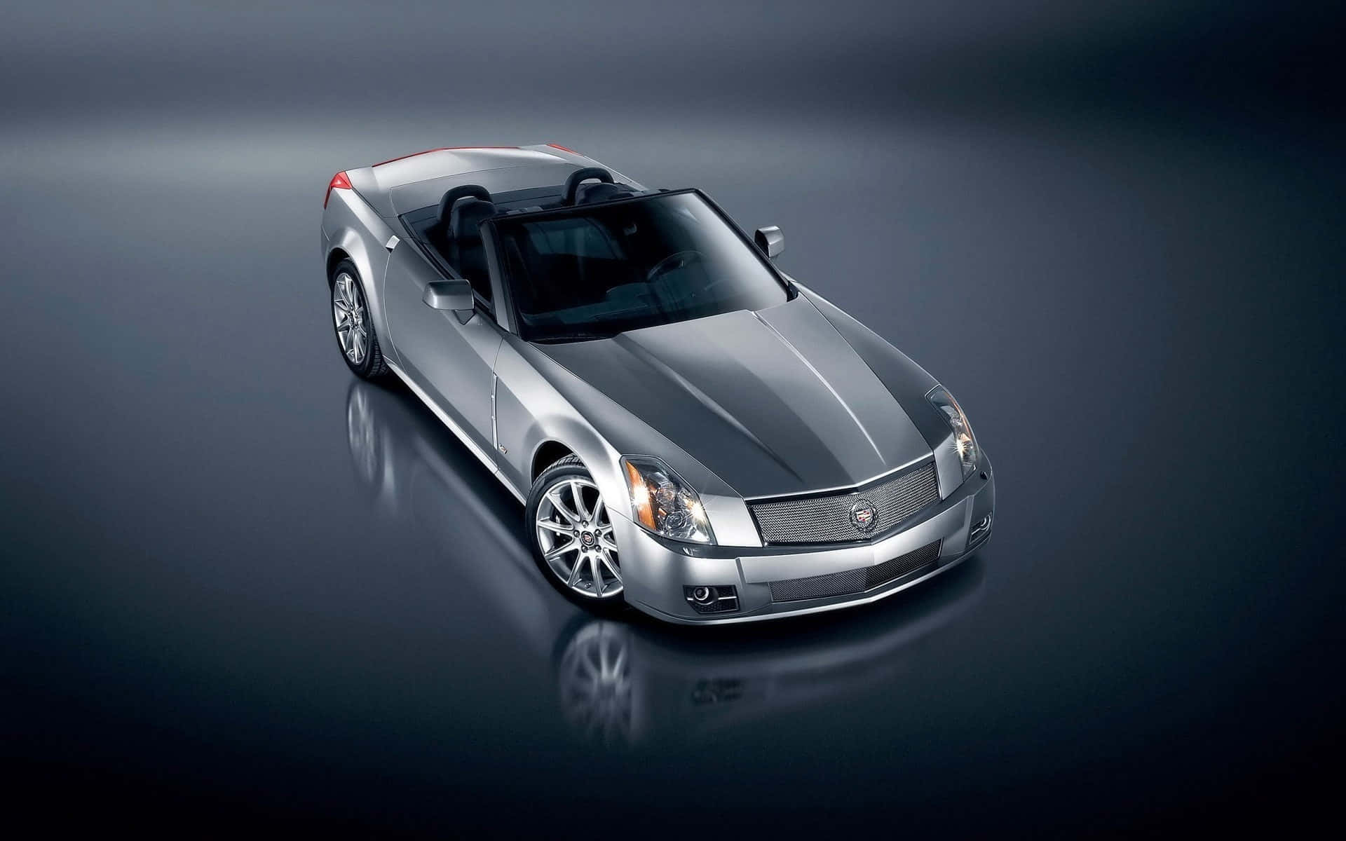 Cadillac Xlr 桌布 壁紙 — 一輛時尚的銀色敞篷跑車，配備黑色軟頂，在戲劇性燈光下展示於反光的深色工作室地板上。