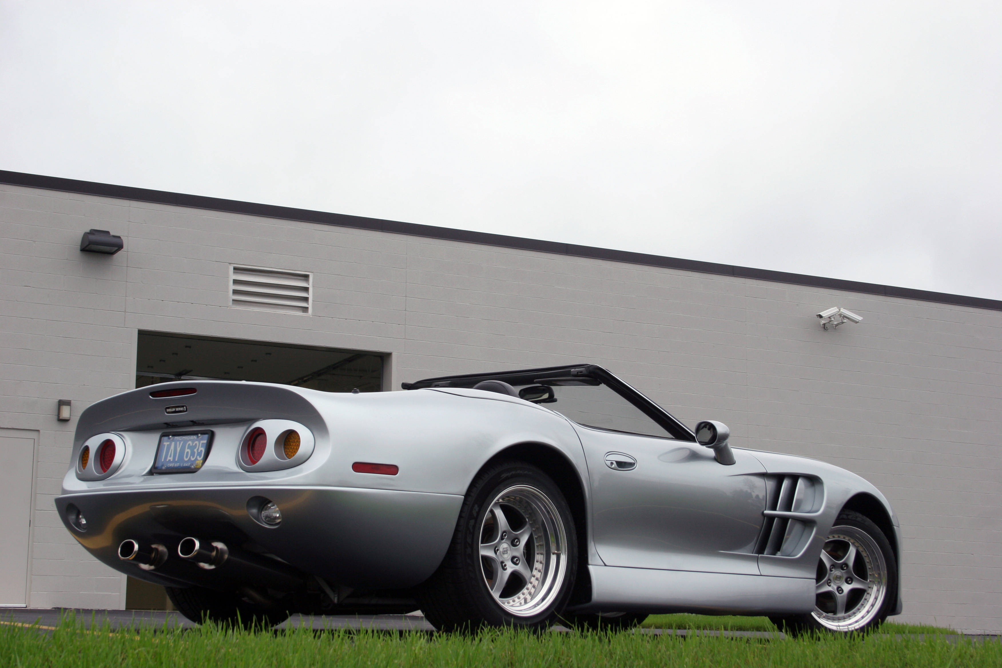 shelby iPhone 桌布 壁紙 — 一輛時尚的銀色敞篷跑車停放在灰色建築物前的草地上，天空多雲。