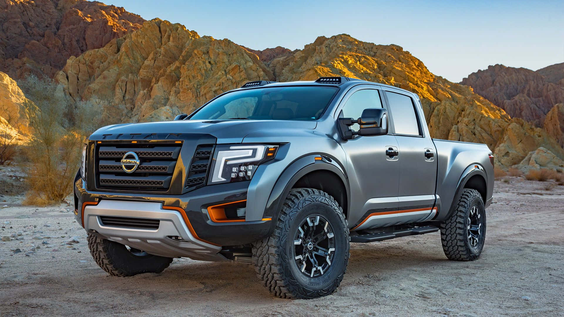 Nissan Titan 桌布 壁紙 — 一輛時尚的銀色日產Frontier皮卡車帶有橙色點綴，在晴朗的藍天下，立於沙質地形上，背景是崎嶇的金色岩層。