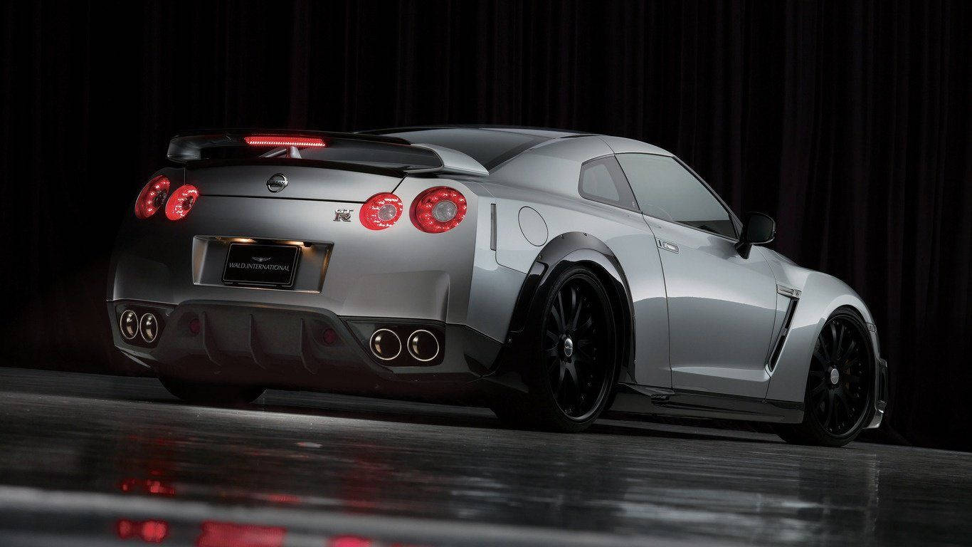 Nissan 桌布 壁紙 — 一輛時尚的銀色日產GT-R跑車，帶有發光的紅色尾燈，在戲劇性的工作室燈光下展示在反光舞台上。
