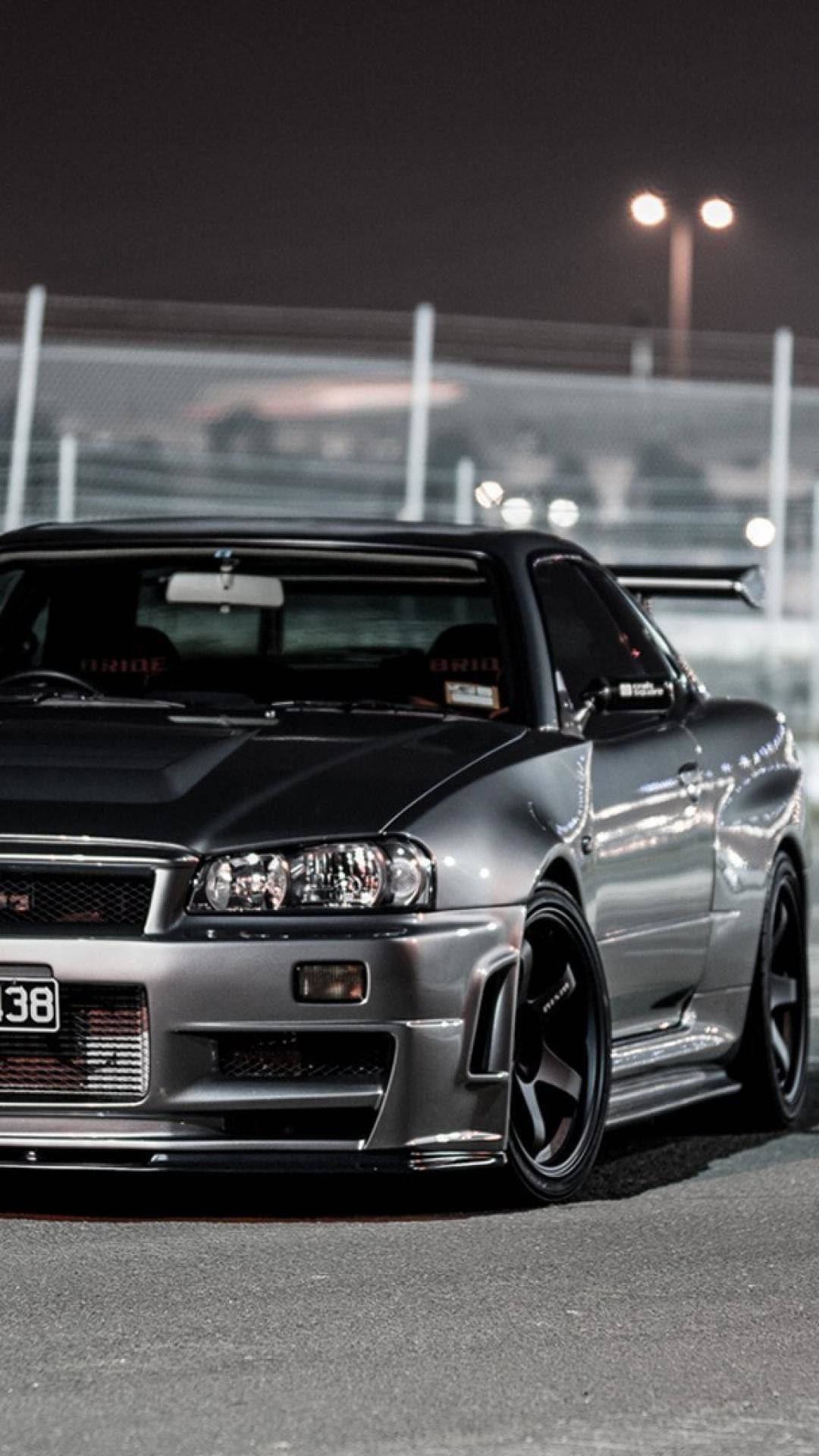 JDM 桌布 壁紙 — 一輛時尚的銀色日產Skyline GT-R在夜間停泊在賽車場上，明亮的泛光燈照射下。