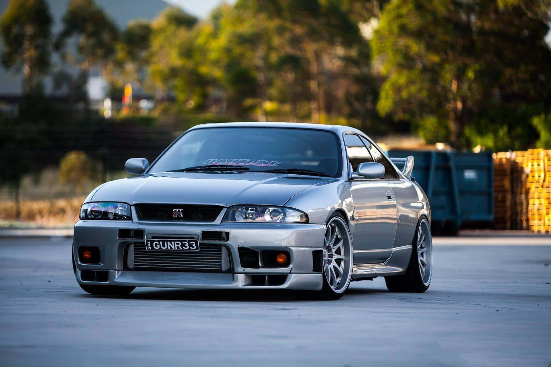Gtr 桌布 壁紙 — 一輛時尚的銀色日產Skyline R34跑車，配備定制輪圈和降低的懸掛系統，在黃金時段停放在混凝土停車場上。