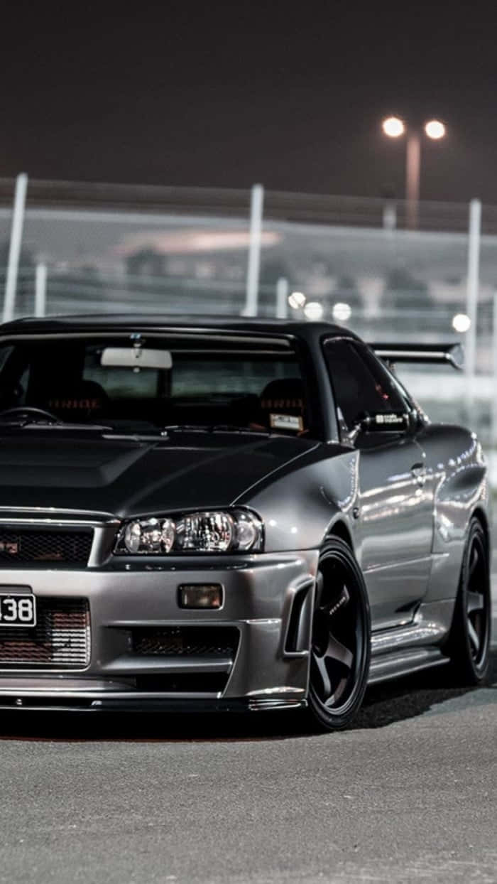 JDM 桌布 壁紙 — 一輛時尚的銀色日產Skyline GT-R在夜間停泊在賽車場上，在明亮的泛光燈下。