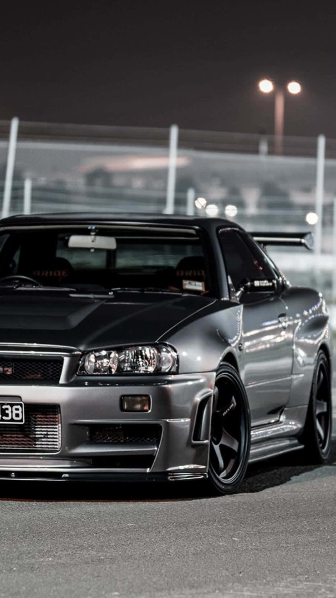 JDM iPhone 桌布 壁紙 — 一輛時尚的銀色日產Skyline GT-R在夜間停泊在賽車場上，明亮的泛光燈照射下。