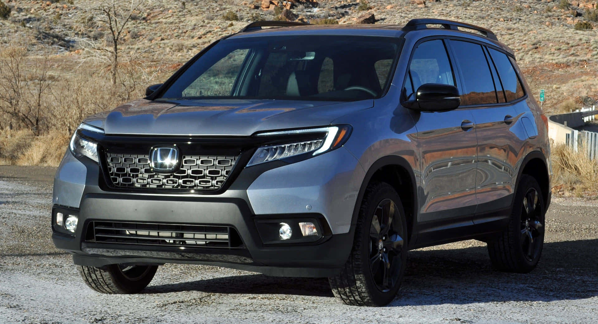 Honda Passport 桌布 壁紙 — 一輛時尚的銀色本田Pilot SUV停在碎石上，背景是崎嶇的地形和乾燥的灌木叢，在明亮的日光下。