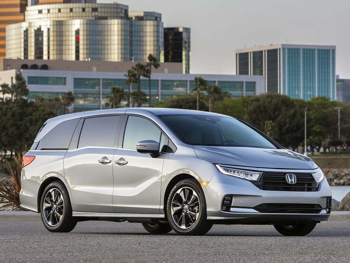 Honda Odyssey 桌布 壁紙 — 一輛時尚的銀色本田小型貨車停在柏油路上，背景有現代玻璃建築和棕櫚樹，在柔和日光下。