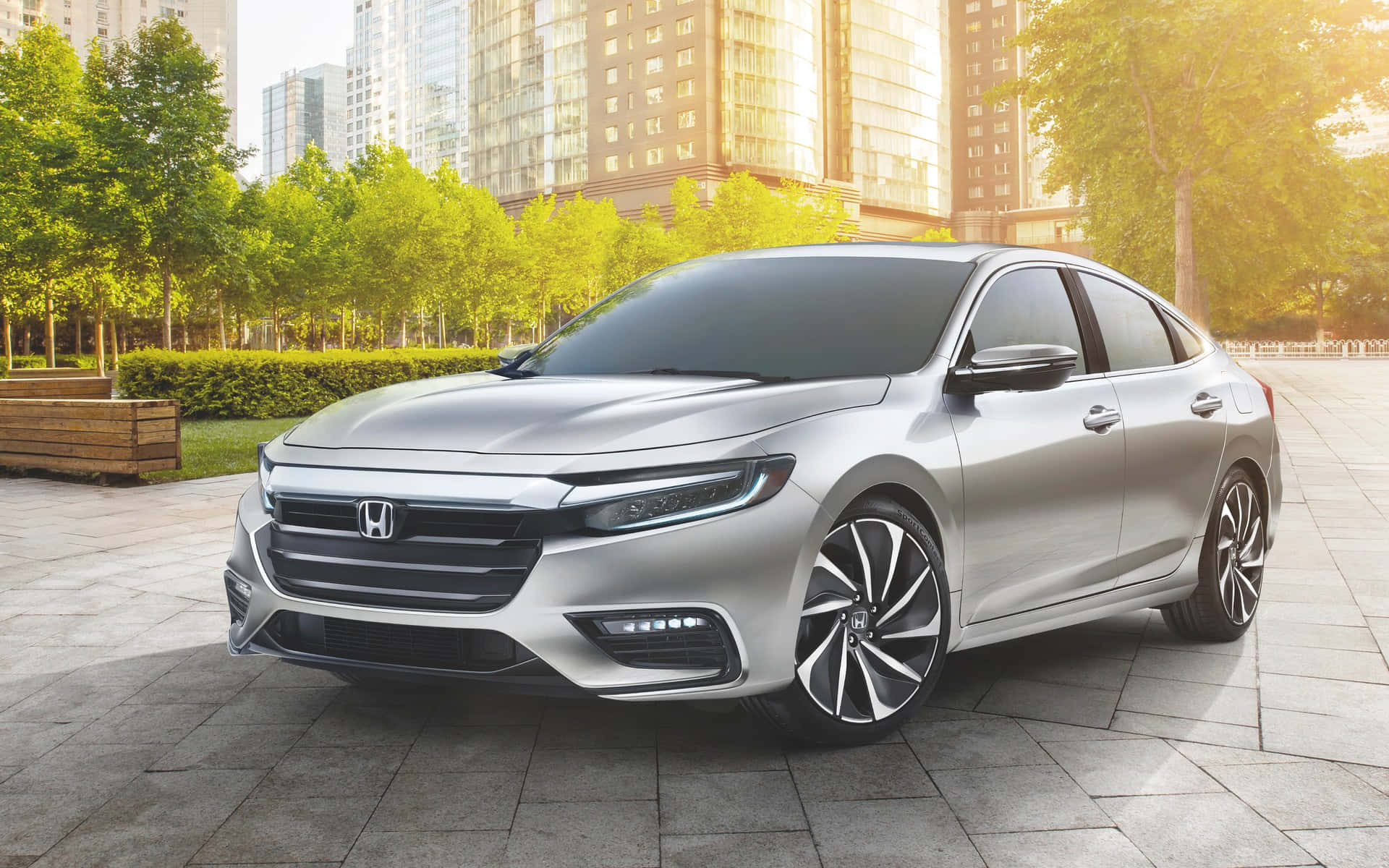 Honda Insight 桌布 壁紙 — 一輛時尚的銀色本田轎車停在石頭廣場上，周圍有綠樹和現代摩天大樓，在金色的午後陽光下閃閃發光。