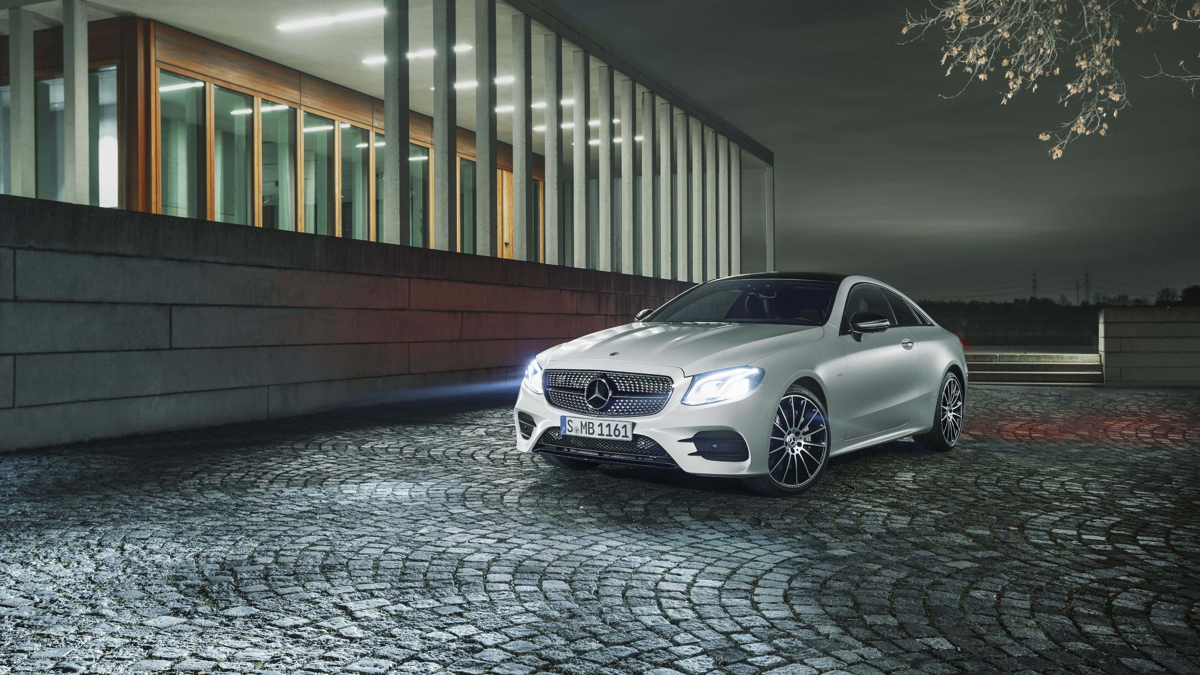 Mercedes Benz Clase E 桌布 壁紙 — 一輛時尚的銀色梅賽德斯E級轎跑車在夜燈下，在現代玻璃建築旁的濕鵝卵石車道上閃閃發光。
