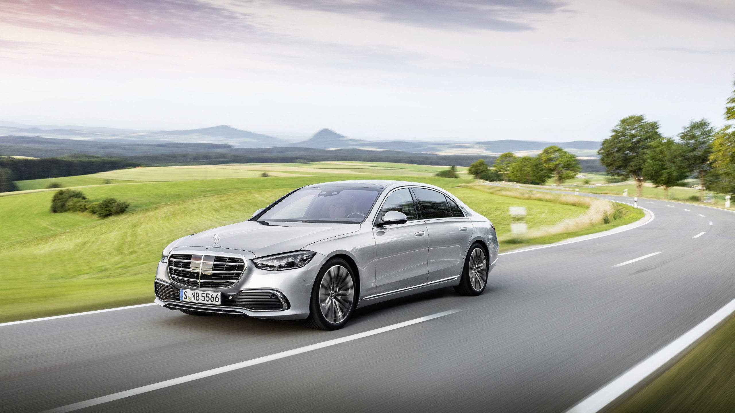 Mercedes 桌布 壁紙 — 一輛時尚的銀色梅賽德斯S級轎車在柔和的暮色天空下，沿著蜿蜒的鄉村道路平穩地滑行，穿過鬱鬱蔥蔥的綠色田野。