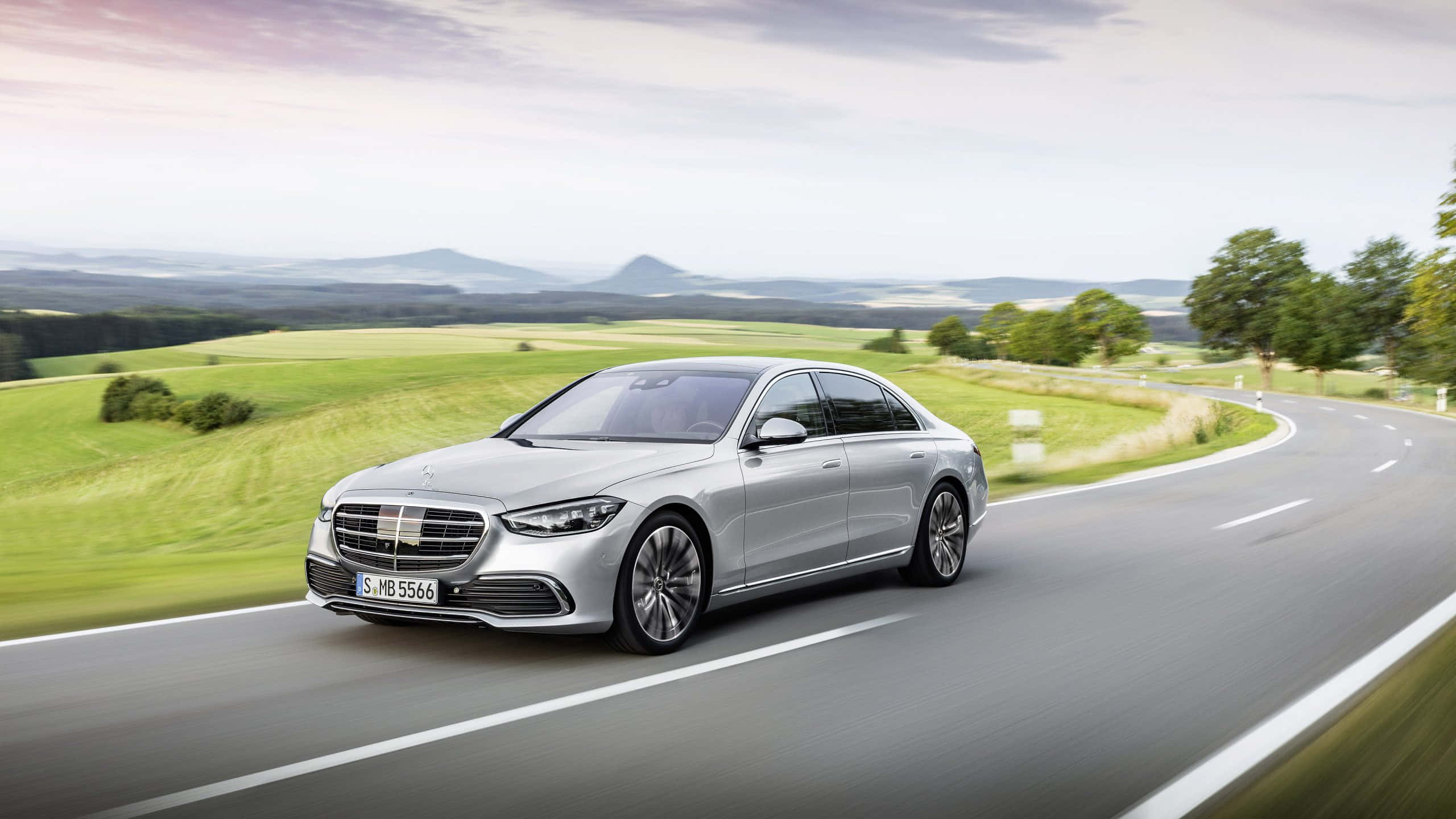 Mercedes 桌布 壁紙 — 一輛時尚的銀色梅賽德斯S級轎車沿著蜿蜒的鄉村道路滑行，穿過茂密的綠色田野，天空柔和多雲。