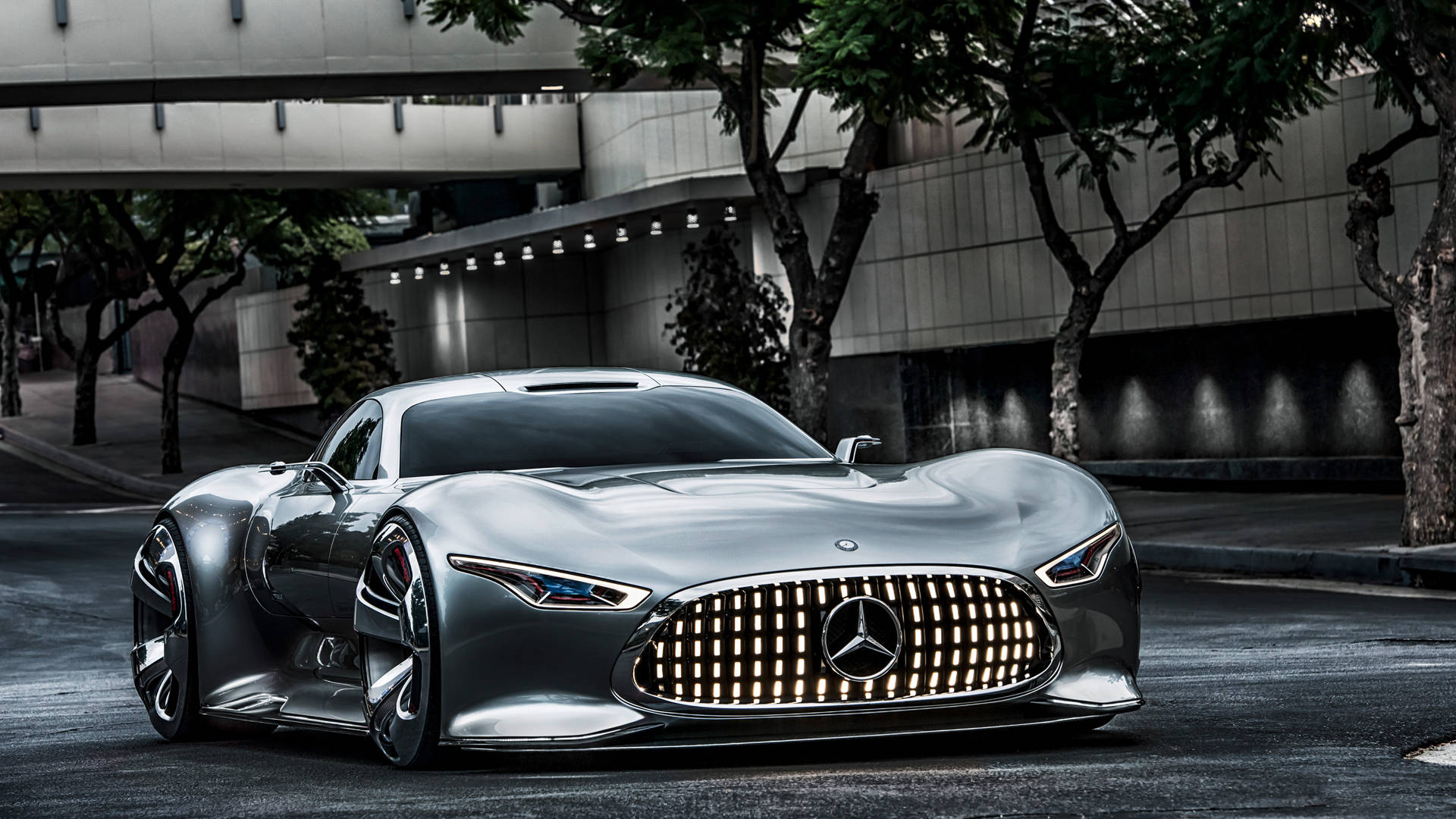 benz 4K 桌布 壁紙 — 一輛時尚的銀色梅賽德斯跑車在都市街燈下閃耀，旁邊是現代建築和枝葉茂盛的樹木。