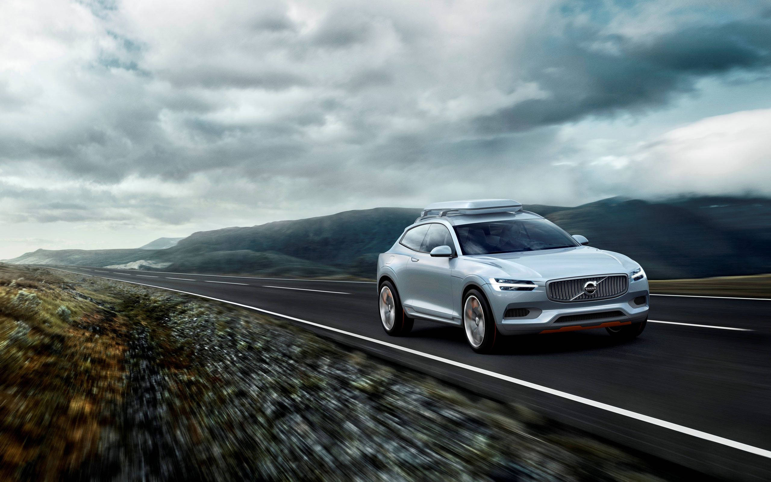 Volvo 桌布 壁紙 — 一輛時尚的銀色沃爾沃SUV在戲劇性的多雲天空下沿著蜿蜒的山路飛馳。