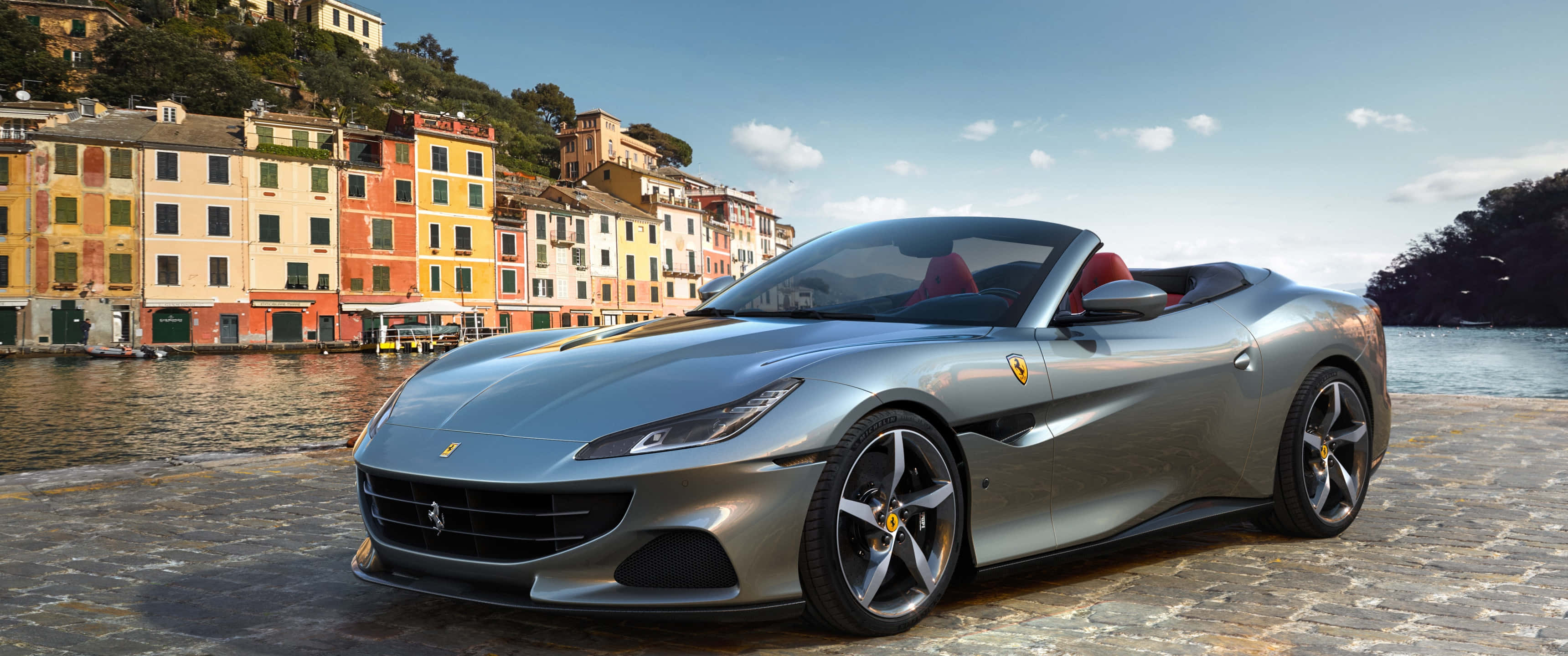 Ferrari Portofino 桌布 壁紙 — 一輛時尚的銀色法拉利Portofino敞篷跑車停泊在鵝卵石海濱，背景是色彩繽紛的義大利建築物和寧靜的水面。
