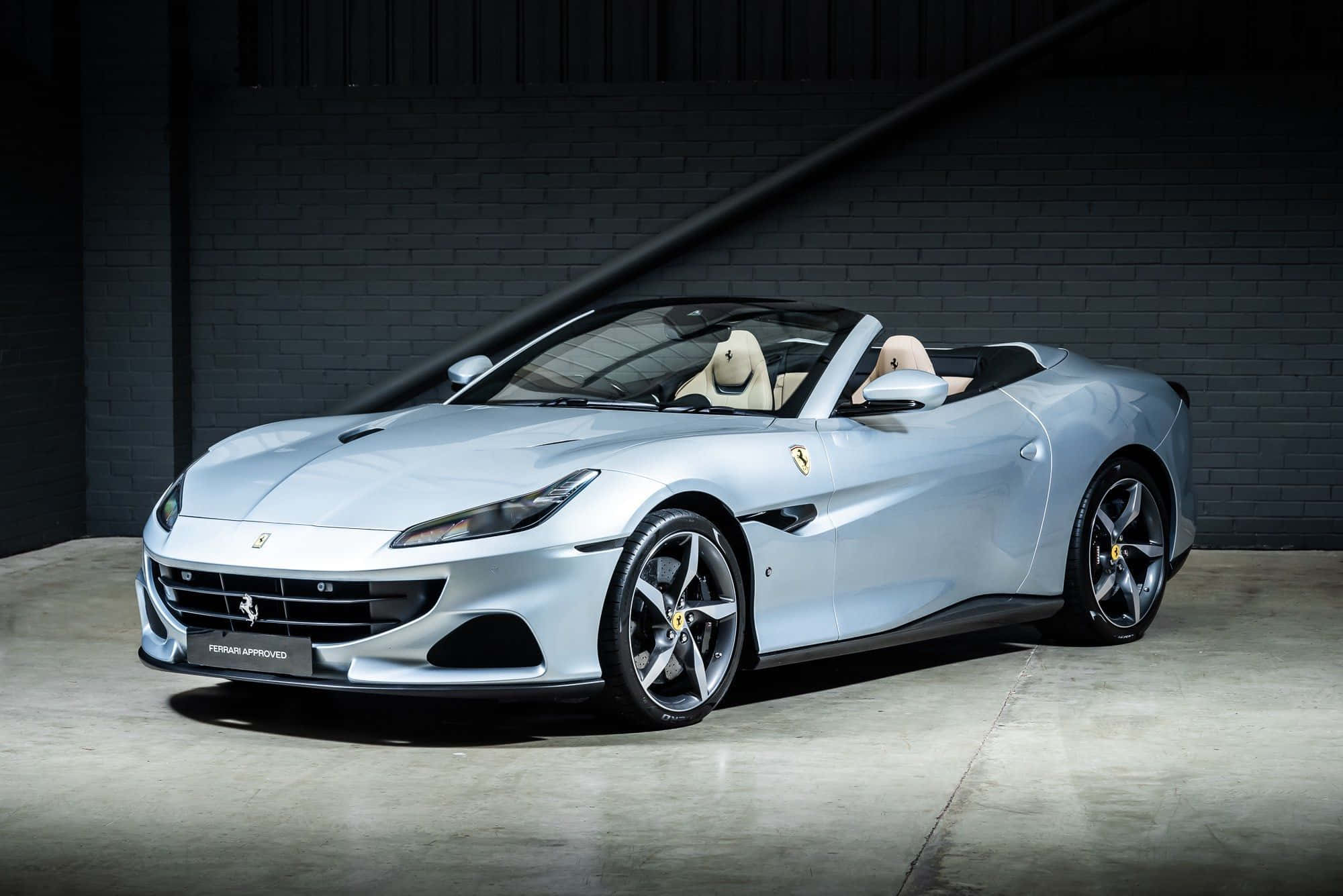 Ferrari Portofino 桌布 壁紙 — 一輛時尚的銀色法拉利Portofino敞篷車，配備米色皮革座椅，停放在光線昏暗的工業車庫中，車庫有裸露的磚牆。