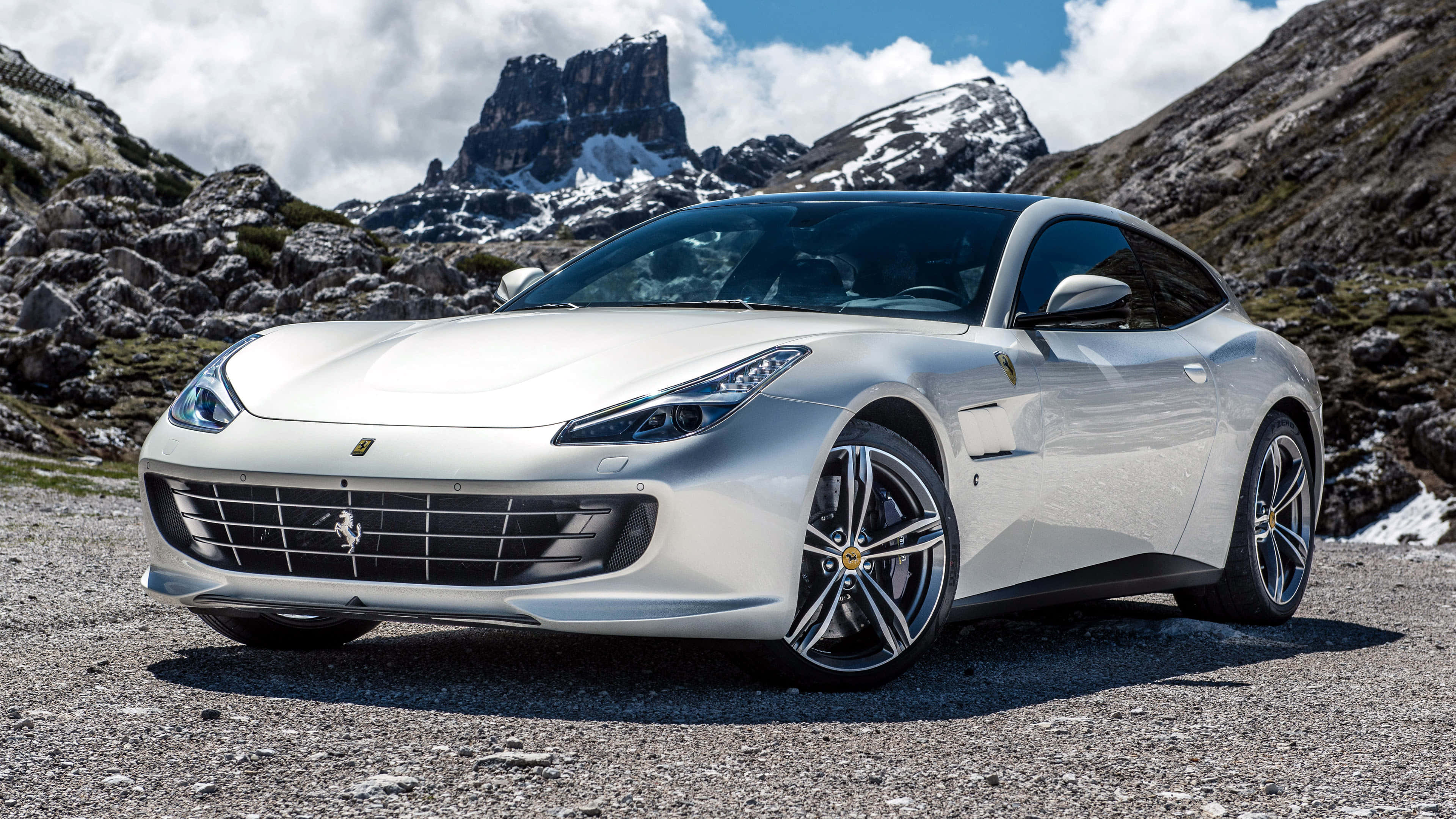 Ferrari Gtc4lusso 桌布 壁紙 — 一輛時尚的銀色法拉利停泊在碎石山路，背景有白雪覆蓋的山峰和岩石地形，天空部分多雲。