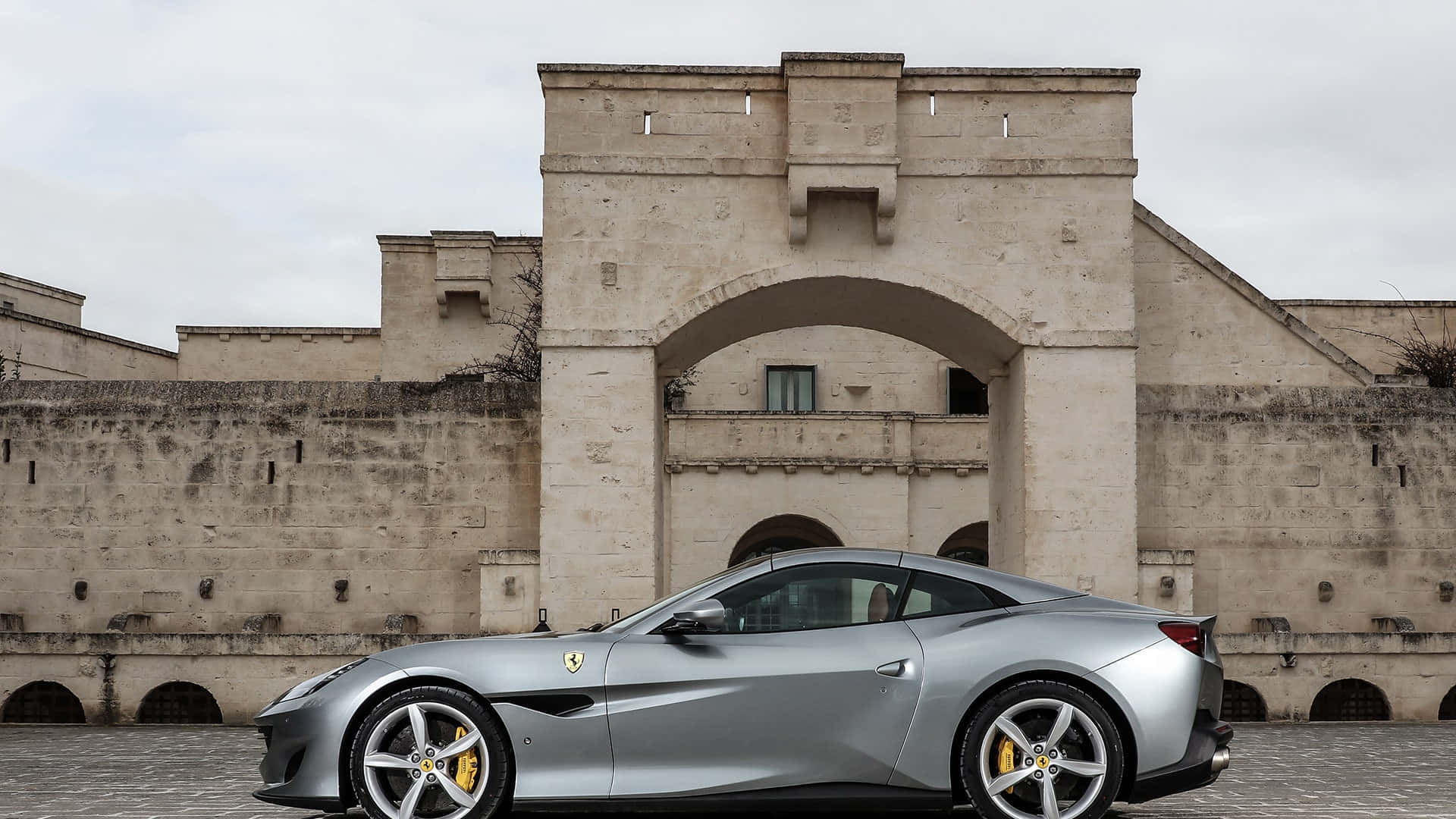 Ferrari Portofino 桌布 壁紙 — 一輛時尚的銀色法拉利跑車停在一座歷史悠久的石頭堡壘前，天空多雲。