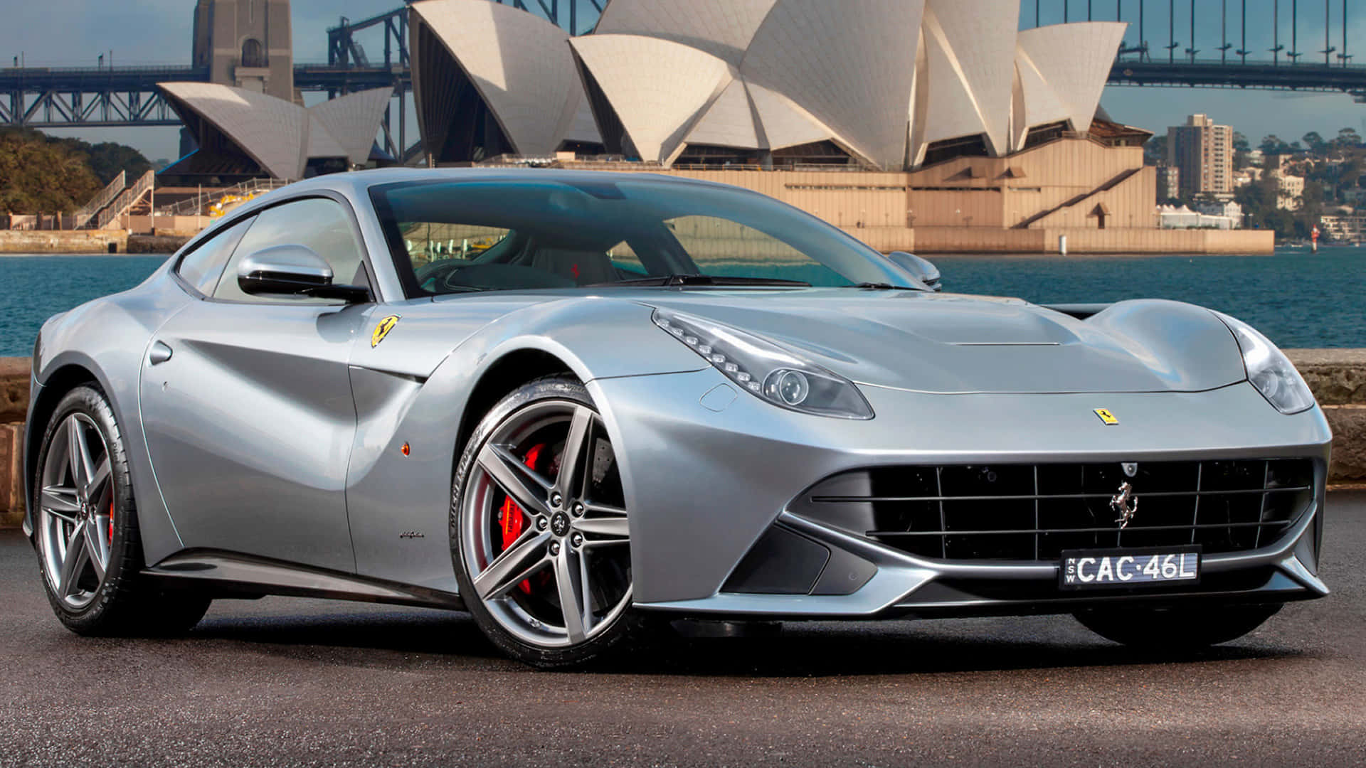 Ferrari F12 Berlinetta 桌布 壁紙 — 一輛時尚的銀色法拉利跑車停在水邊，背景是標誌性的雪梨歌劇院和港灣大橋。