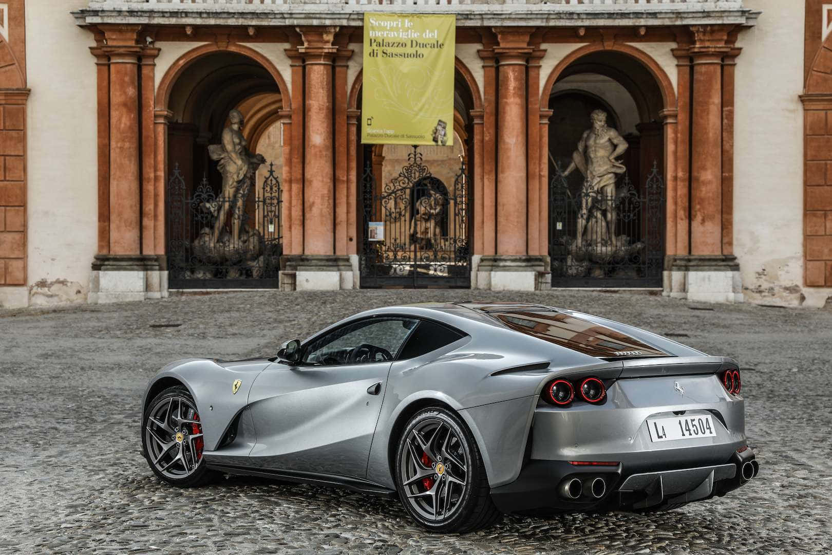 Ferrari 812 Superfast 桌布 壁紙 — 一輛時尚的銀色法拉利跑車停泊在鵝卵石路上，前方是擁有雕像和拱門入口的宏偉歷史宮殿。