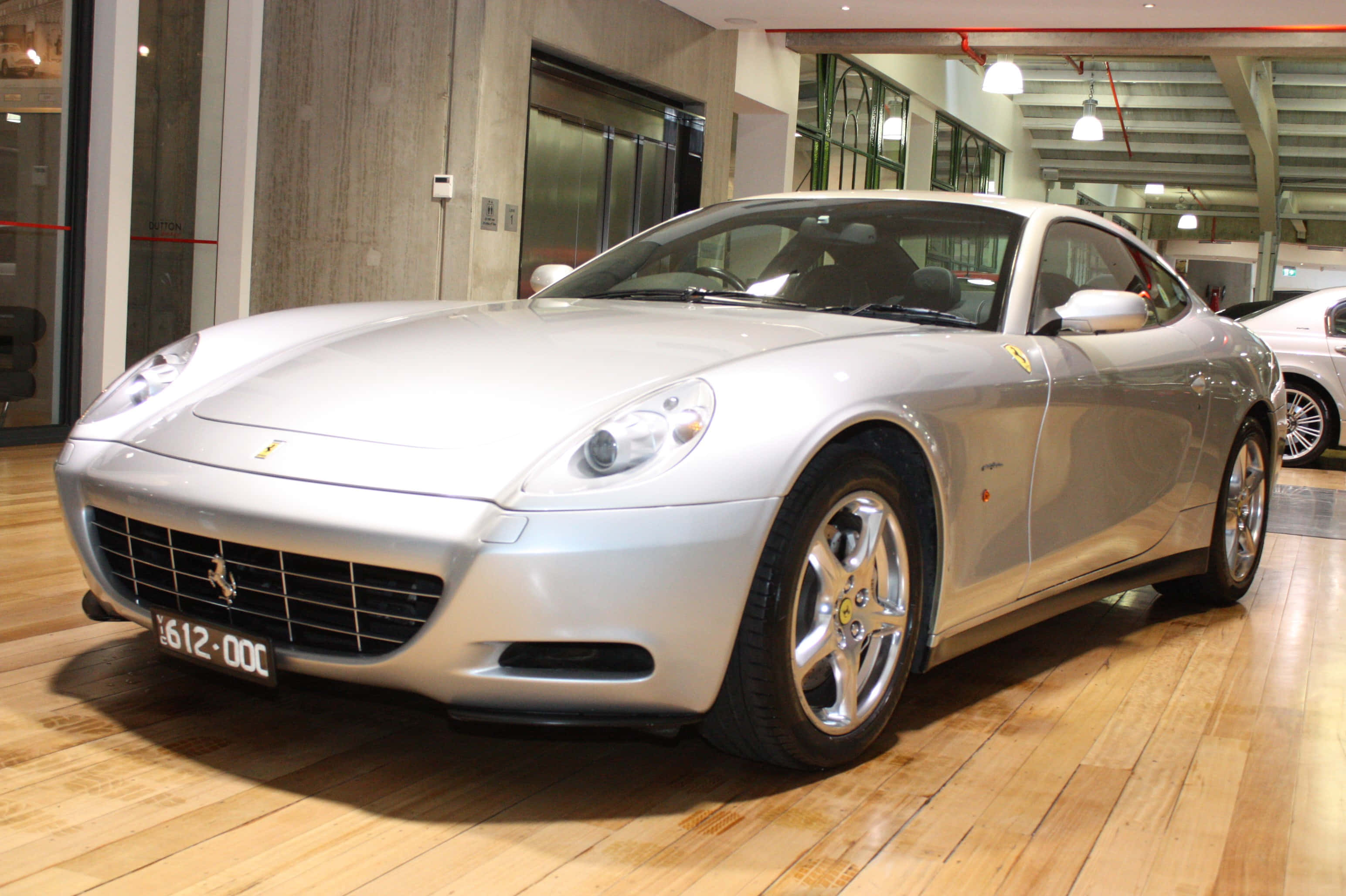 Ferrari 612 Scaglietti 桌布 壁紙 — 一輛時尚的銀色法拉利跑車在展廳燈光下閃耀，停放在拋光木地板上。