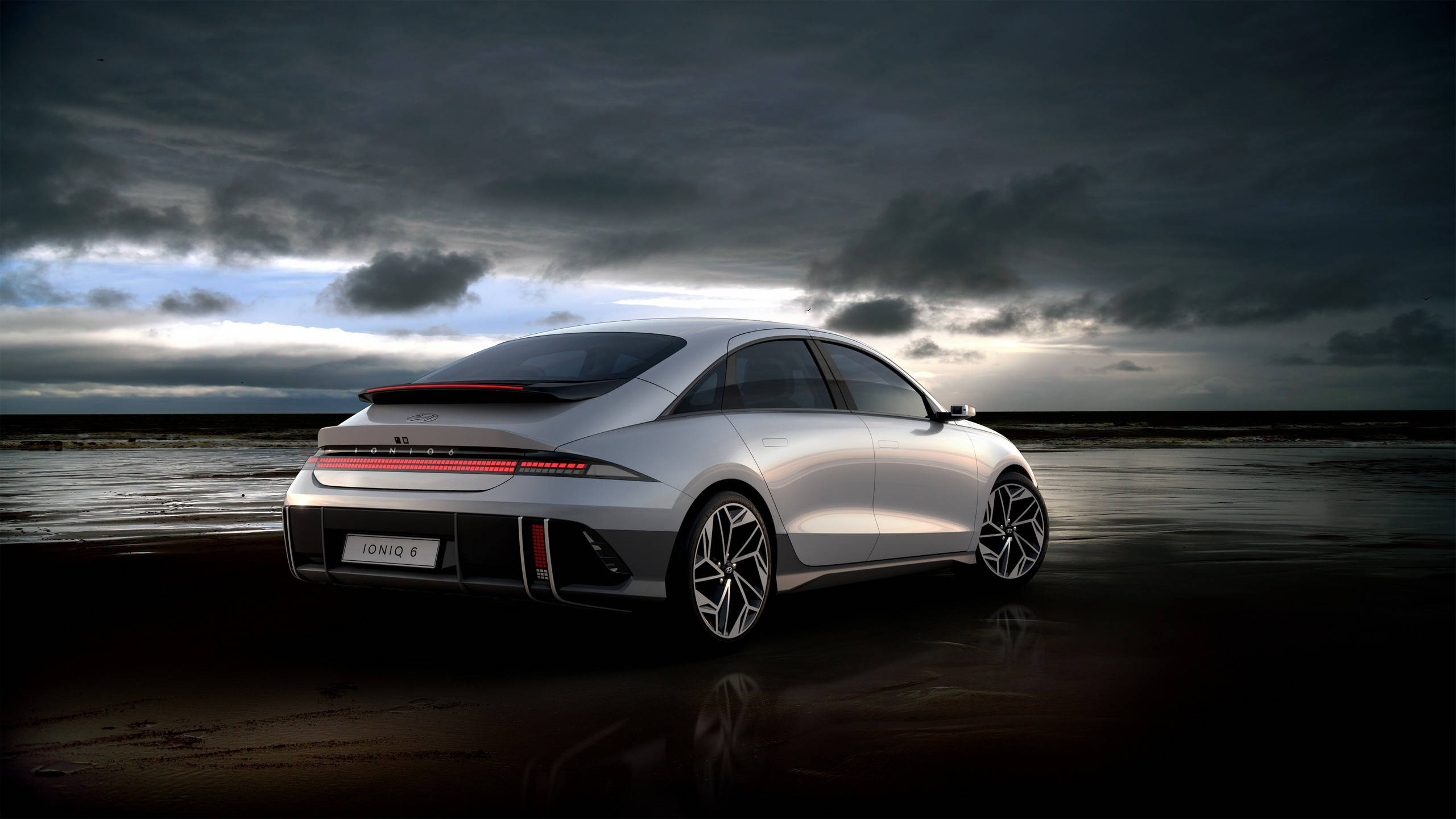 cars 桌布 壁紙 — 一輛時尚的銀色現代 IONIQ 6 電動車停在濕漉漉的海灘上，背景是戲劇性的暴風雨天空，尾燈閃閃發光。