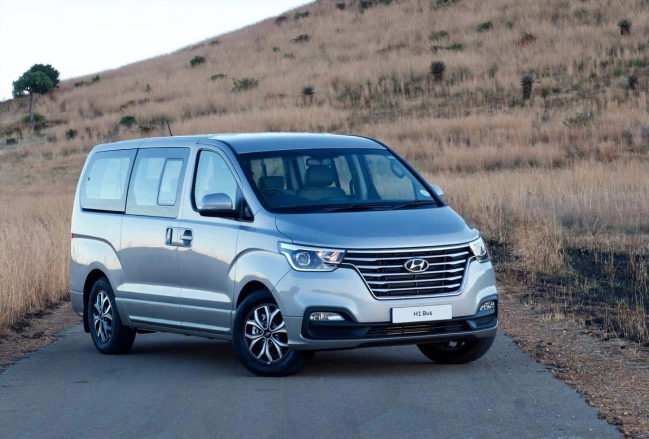 Hyundai 桌布 壁紙 — 一輛時尚的銀色現代HI Bus客貨車停泊在鋪砌道路上，背景是乾燥的草坡，在柔和日光下。