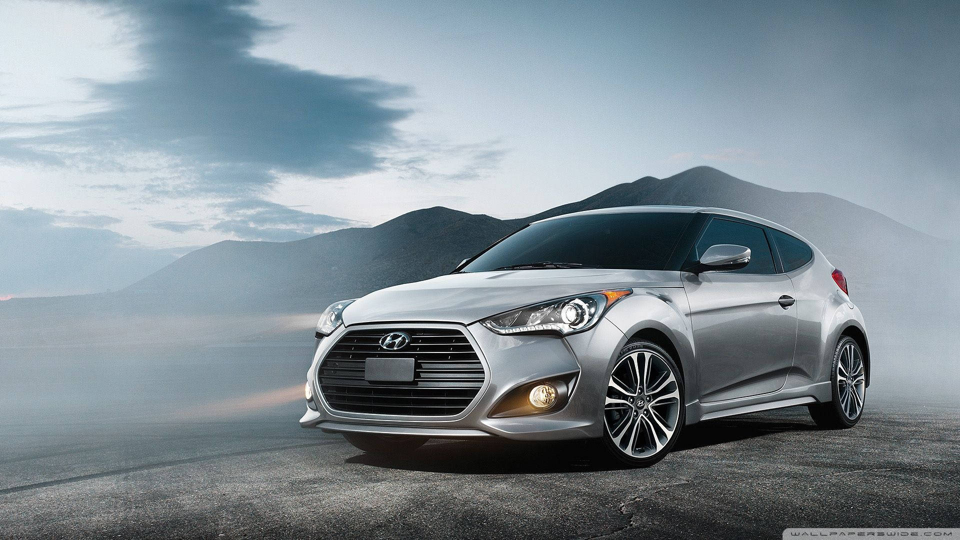 Hyundai 桌布 壁紙 — 一輛時尚的銀色現代Veloster跑車停泊在霧氣瀰漫的道路上，背景是山巒，天空多雲。