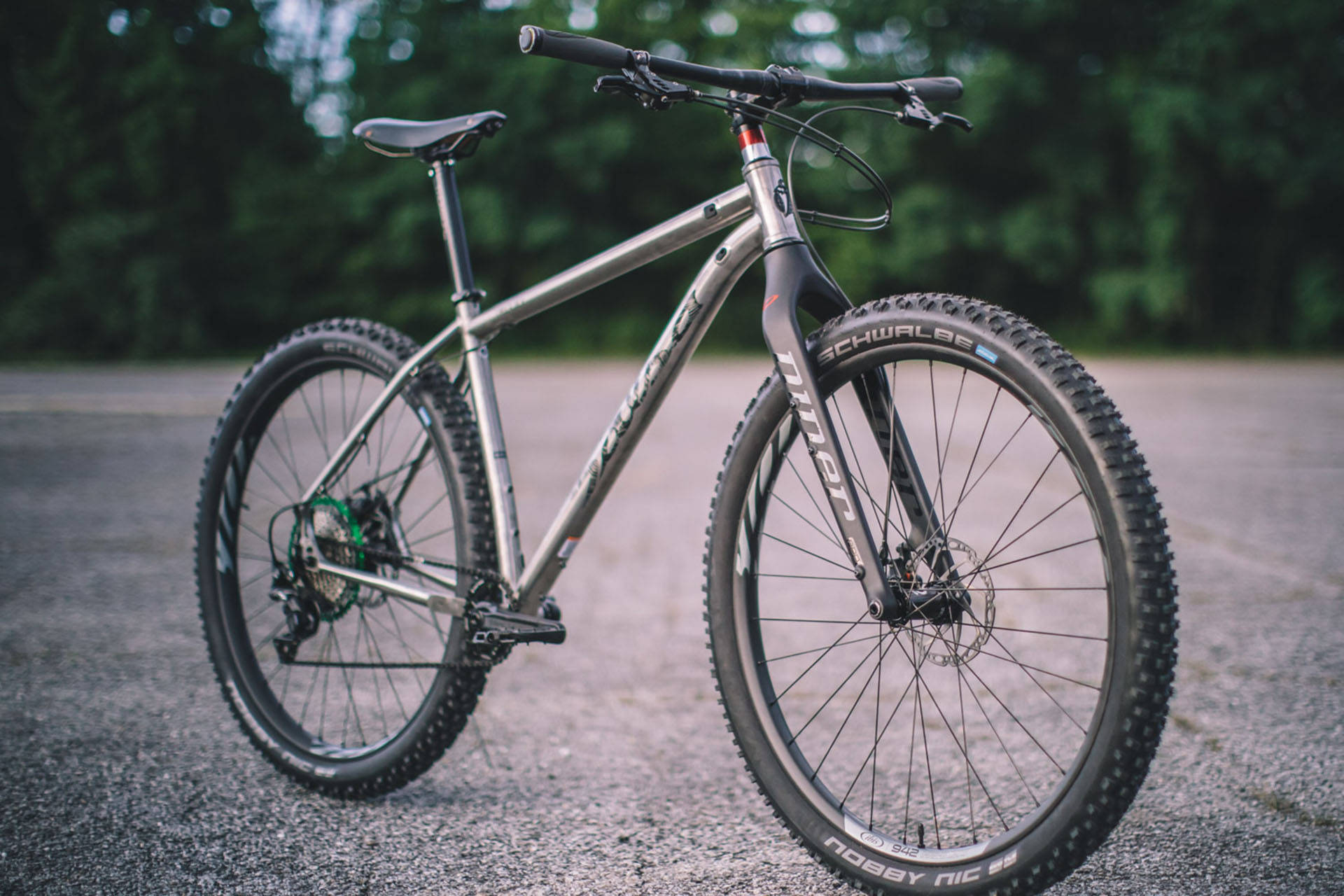 Mountain Bike 桌布 壁紙 — 一輛時尚的銀色登山車，配備厚輪胎，停在碎石路上，周圍是茂密的綠樹。