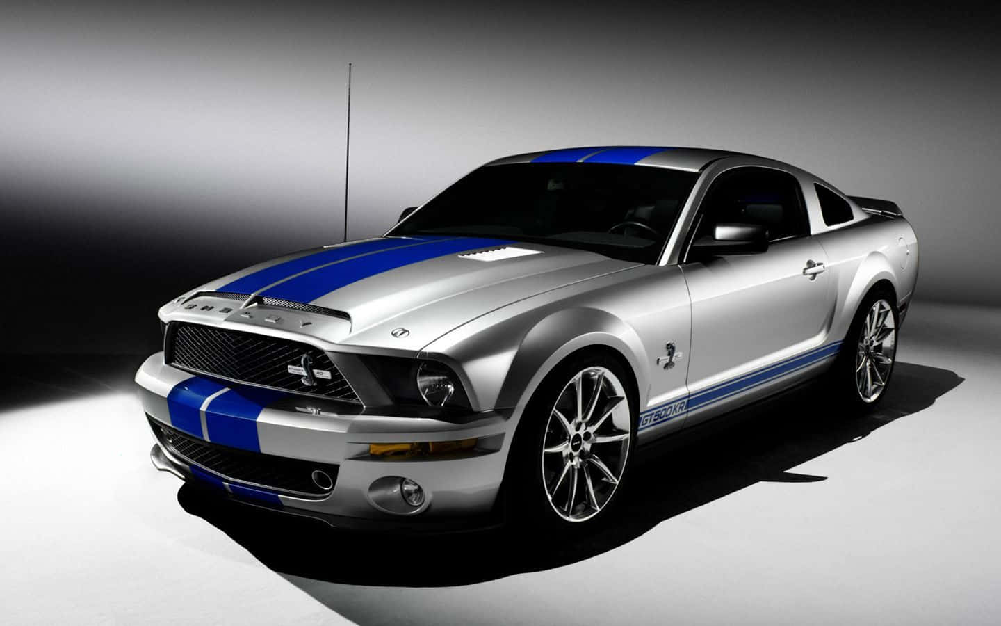 肌肉車 桌布 壁紙 — 一輛時尚的銀色福特野馬 Shelby GT500，帶有醒目的藍色賽車條紋，在戲劇性的工作室燈光下閃閃發光。