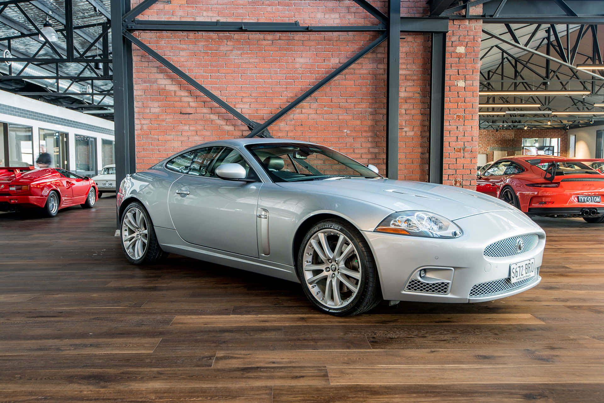 Jaguar Xkr 桌布 壁紙 — 一輛時尚的銀色積架XK轎跑車在明亮的燈光下閃閃發光，位於質樸的磚木汽車展廳，背景有紅色跑車。