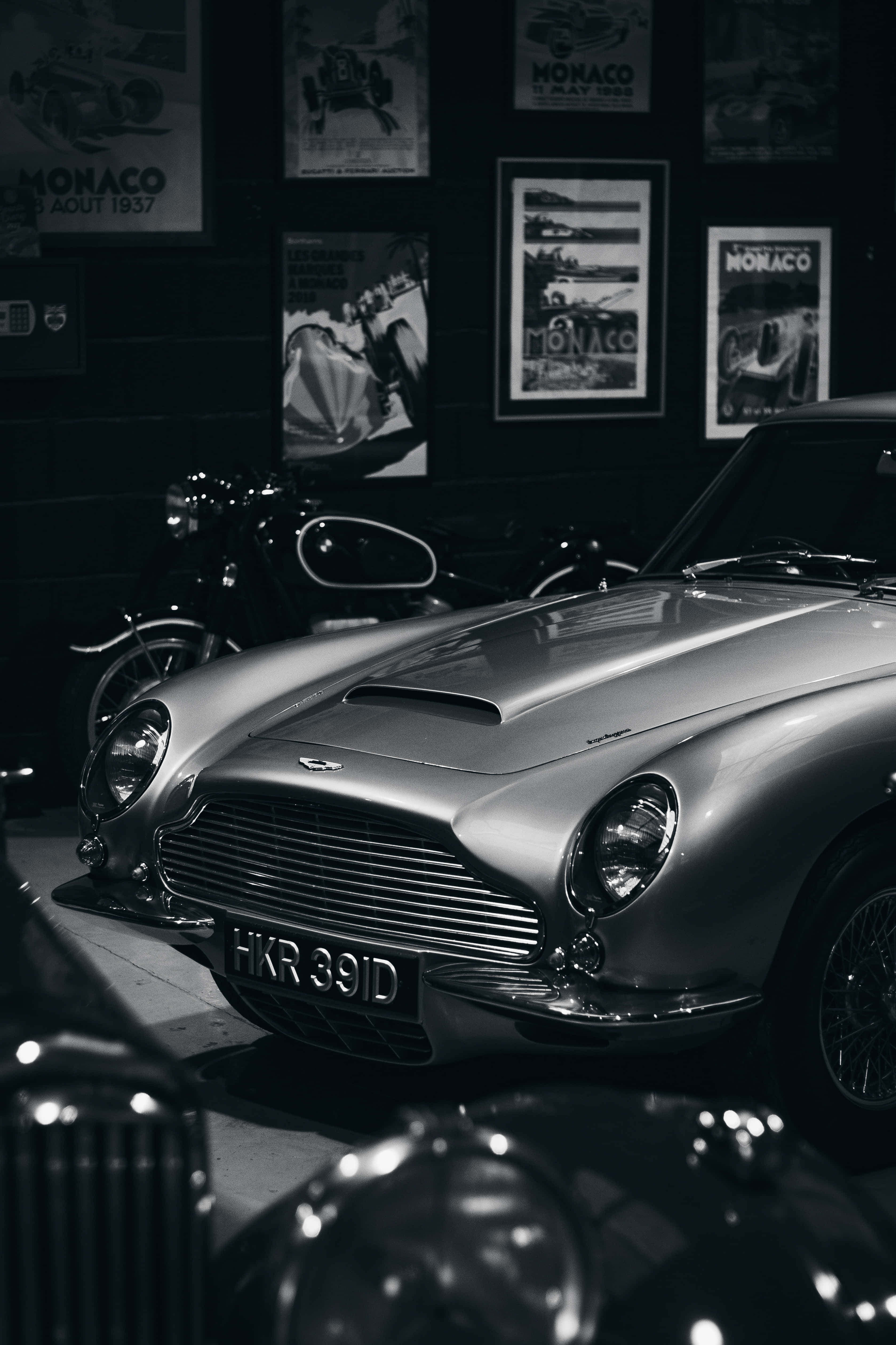 Vintage Ferrari 桌布 壁紙 — 一輛時尚的銀色經典 Aston Martin DB5 在昏暗燈光下閃耀，位於裝飾有摩納哥賽車海報的復古汽車博物館中。