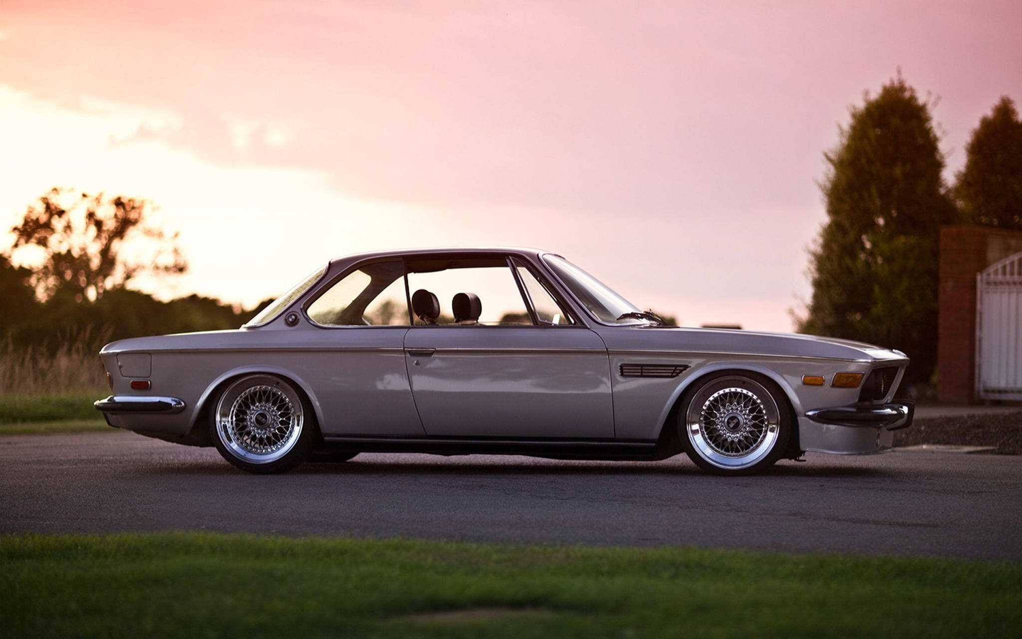 classic bmw 桌布 壁紙 — 一輛時尚的銀色經典BMW雙門轎跑車，配備鋼絲輪圈，停泊在車道上，夕陽西下，天空粉紅，草地翠綠。