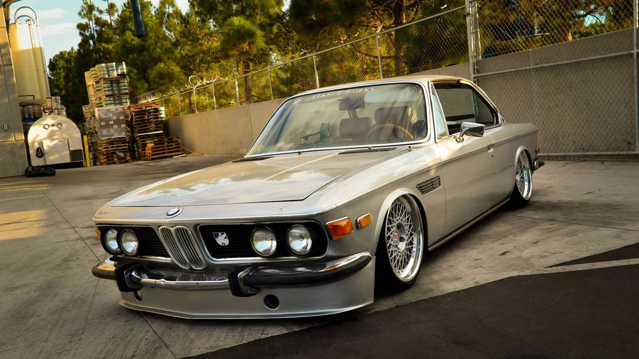 classic bmw 桌布 壁紙 — 一輛時尚的銀色經典寶馬轎跑車，配備定制輪圈，停在混凝土上，靠近鐵絲網圍欄和樹木。