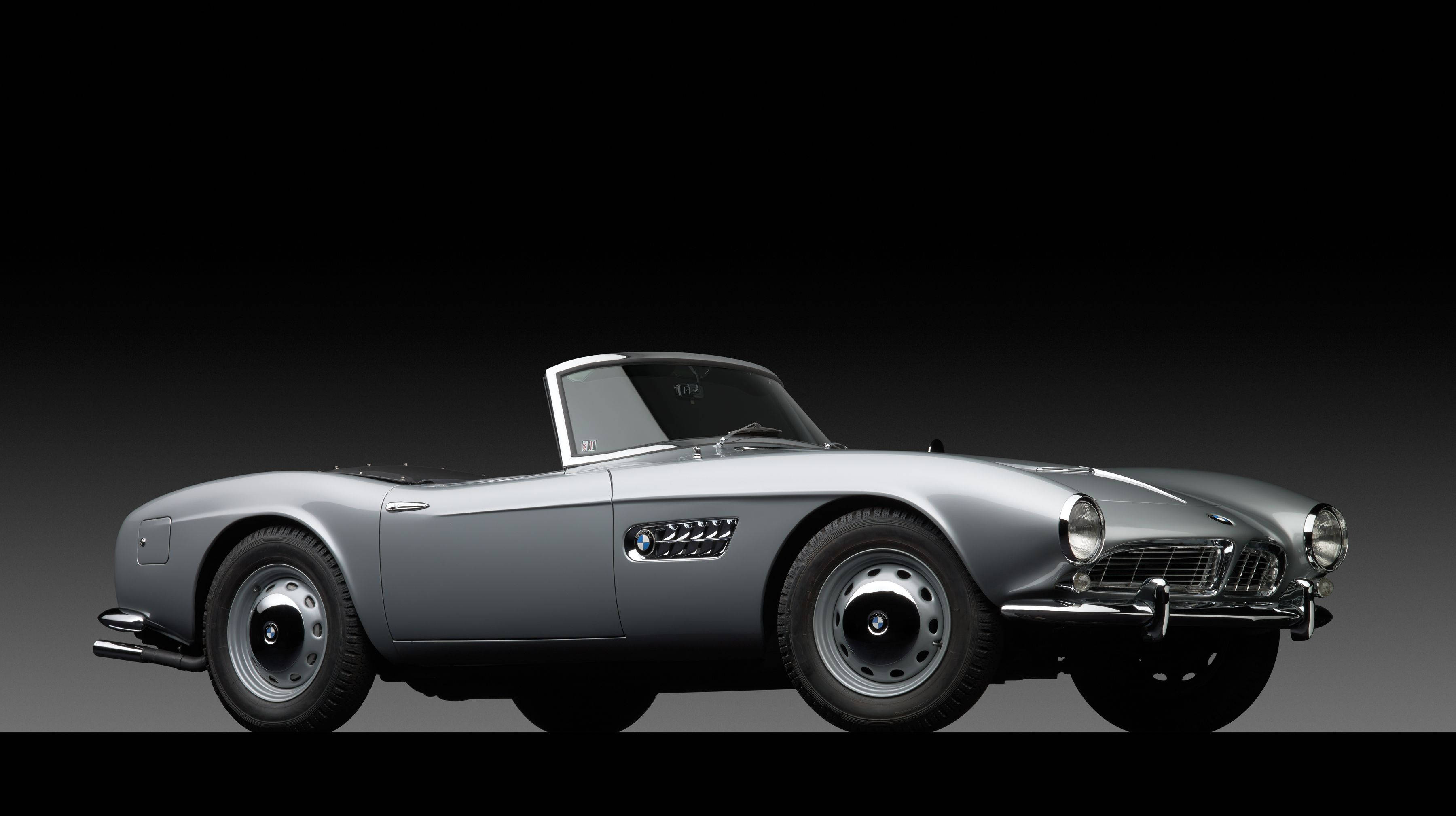 classic bmw 桌布 壁紙 — 一輛時尚的銀色經典敞篷跑車，帶有鍍鉻飾邊和圓形頭燈，在深色漸變背景前矗立。
