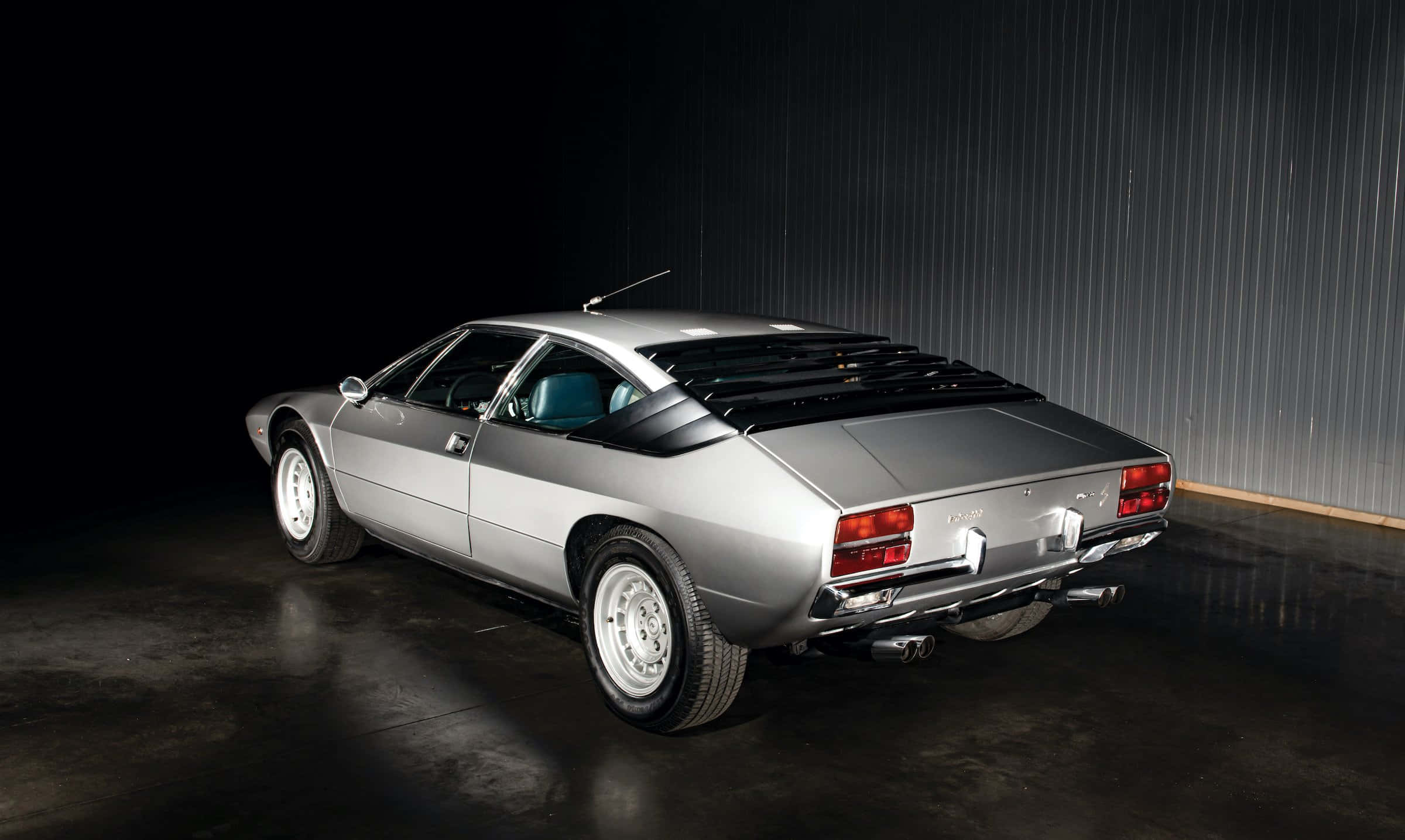 Lamborghini Urraco 桌布 壁紙 — 一輛時尚的銀色經典跑車，配備黑色車頂和後通風口，在光線昏暗的工業車庫中展示，車庫牆壁為波紋狀。