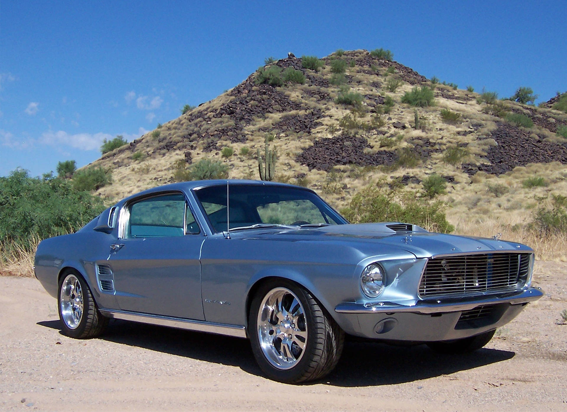 shelby iPhone 桌布 壁紙 — 一輛時尚的銀色經典野馬跑車停在塵土飛揚的沙漠公路上，背景是崎嶇的山丘和仙人掌，天空湛藍。